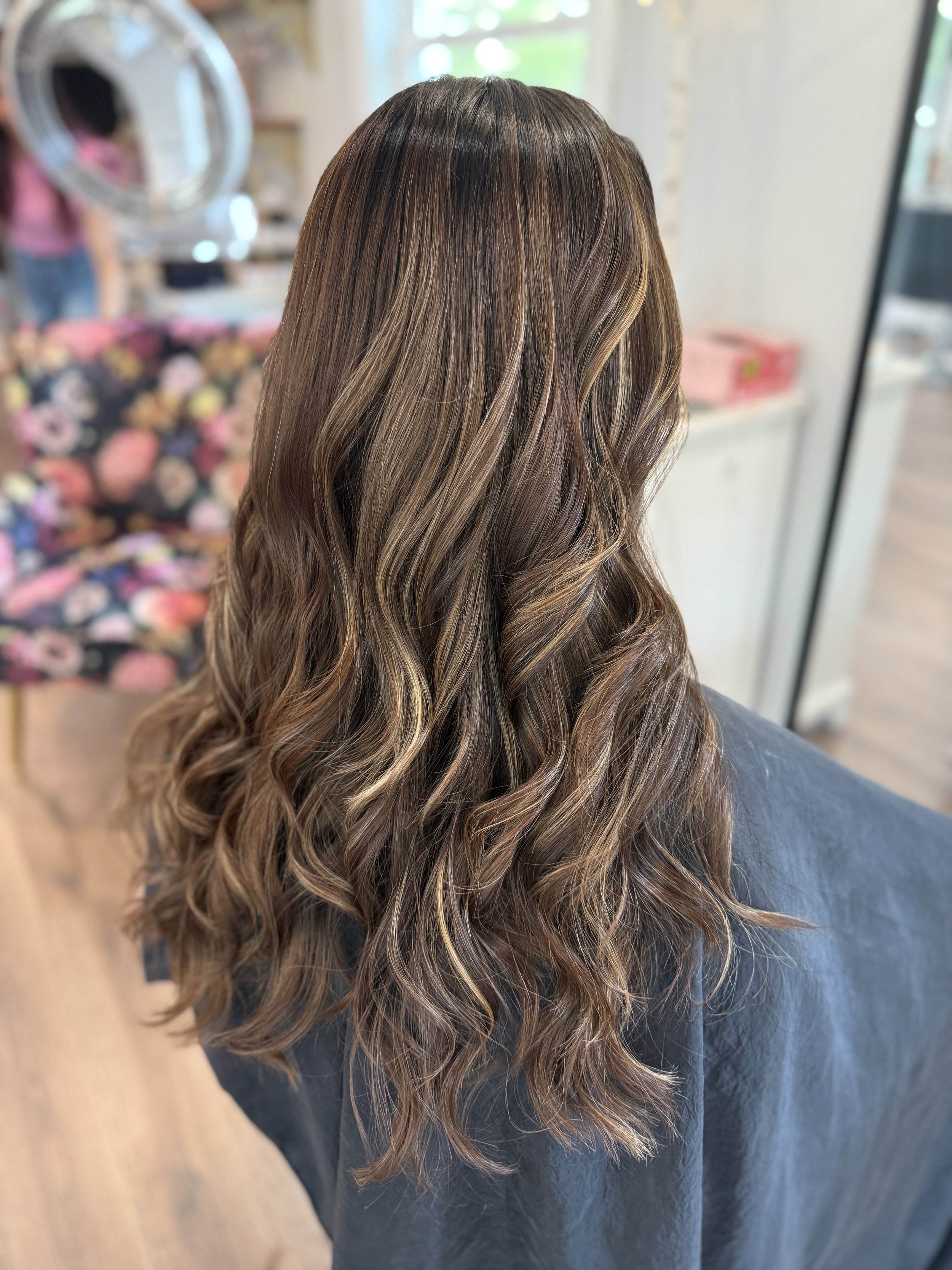 Woman with long, wavy, brown hair with blonde highlights sitting in a salon chair.