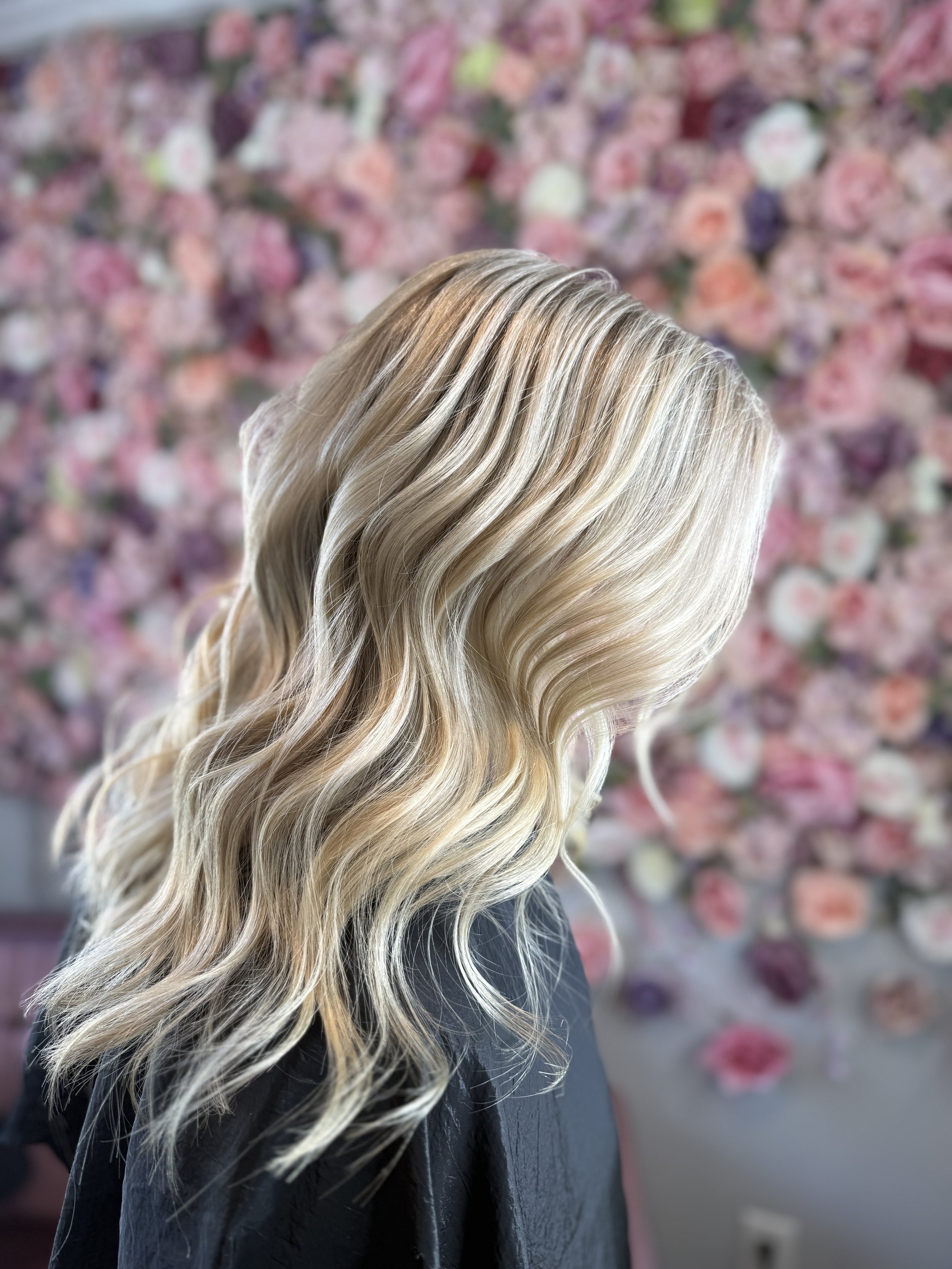 Woman with long, wavy blonde hair, seen from the side, in front of a backdrop of pink and purple flowers.