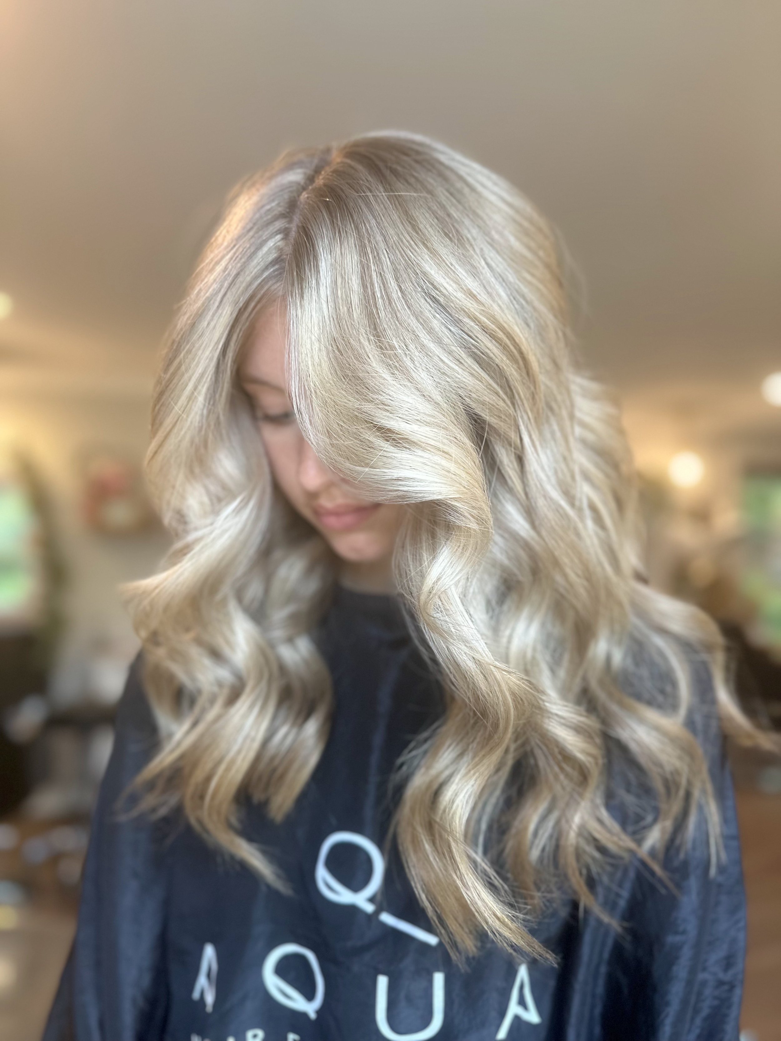 A woman with long, wavy blonde hair in a salon chair, wearing a black cape with white text.