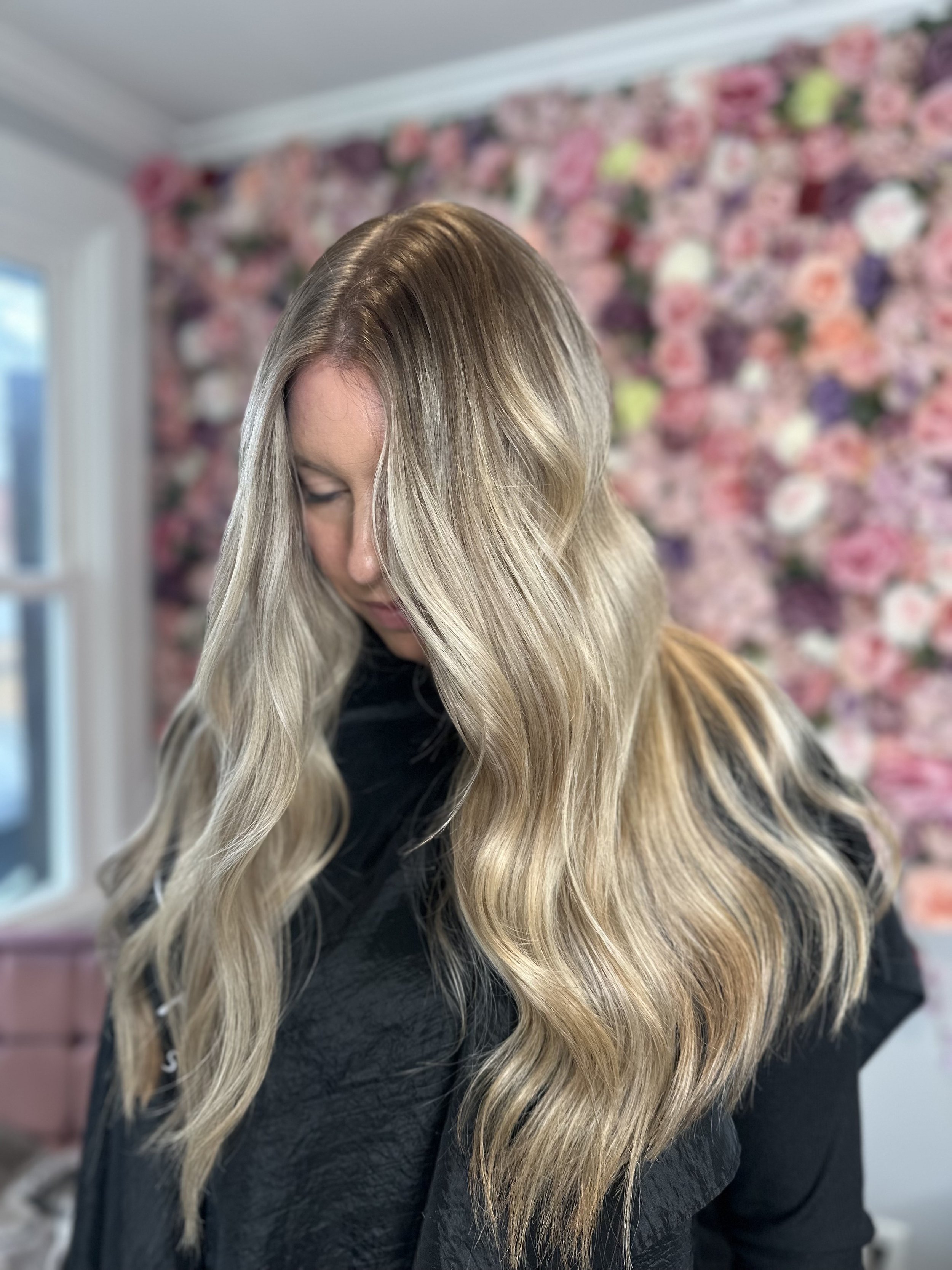 Woman with long, wavy blonde hair in front of a pink floral wall.