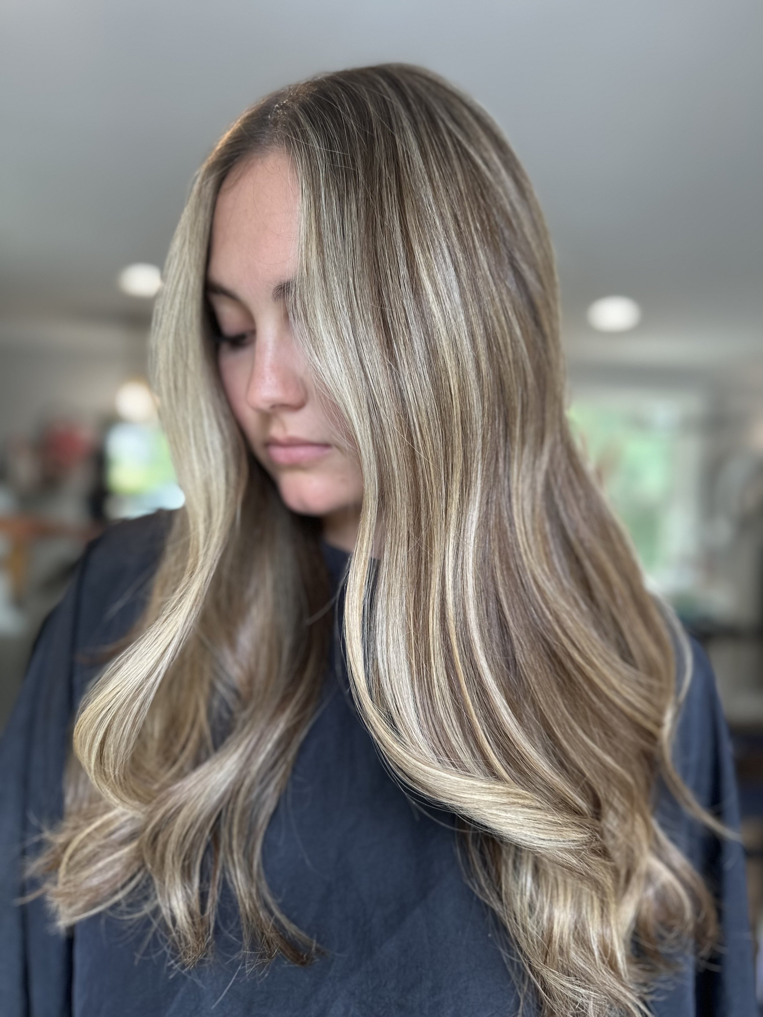A woman with long, styled hair featuring blonde highlights and soft waves, looking down with her eyes closed.