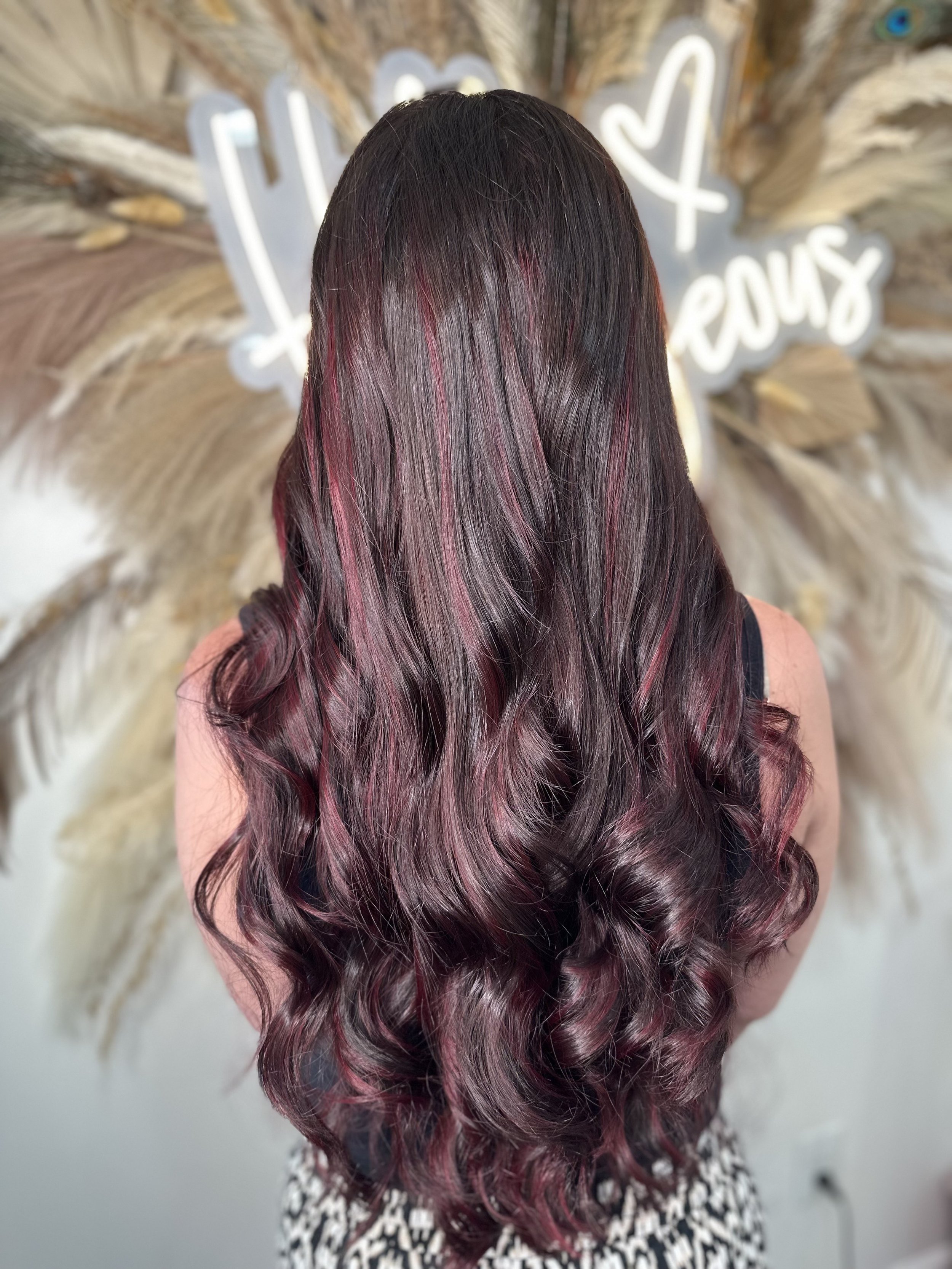 Woman with long, wavy hair dyed in shades of dark purple and burgundy. The background includes a decorative wall with dried grass and a neon sign that says "Love Yourself" with a heart symbol.