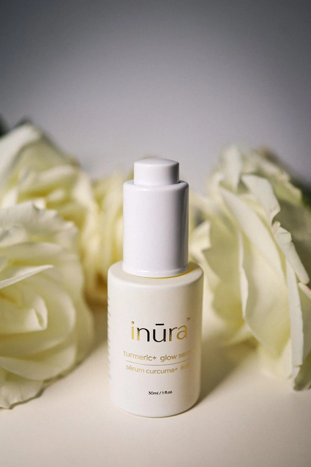 A white bottle of inūra turmeric+ glow skincare serum with a pump dispenser, surrounded by white roses, with gray background.