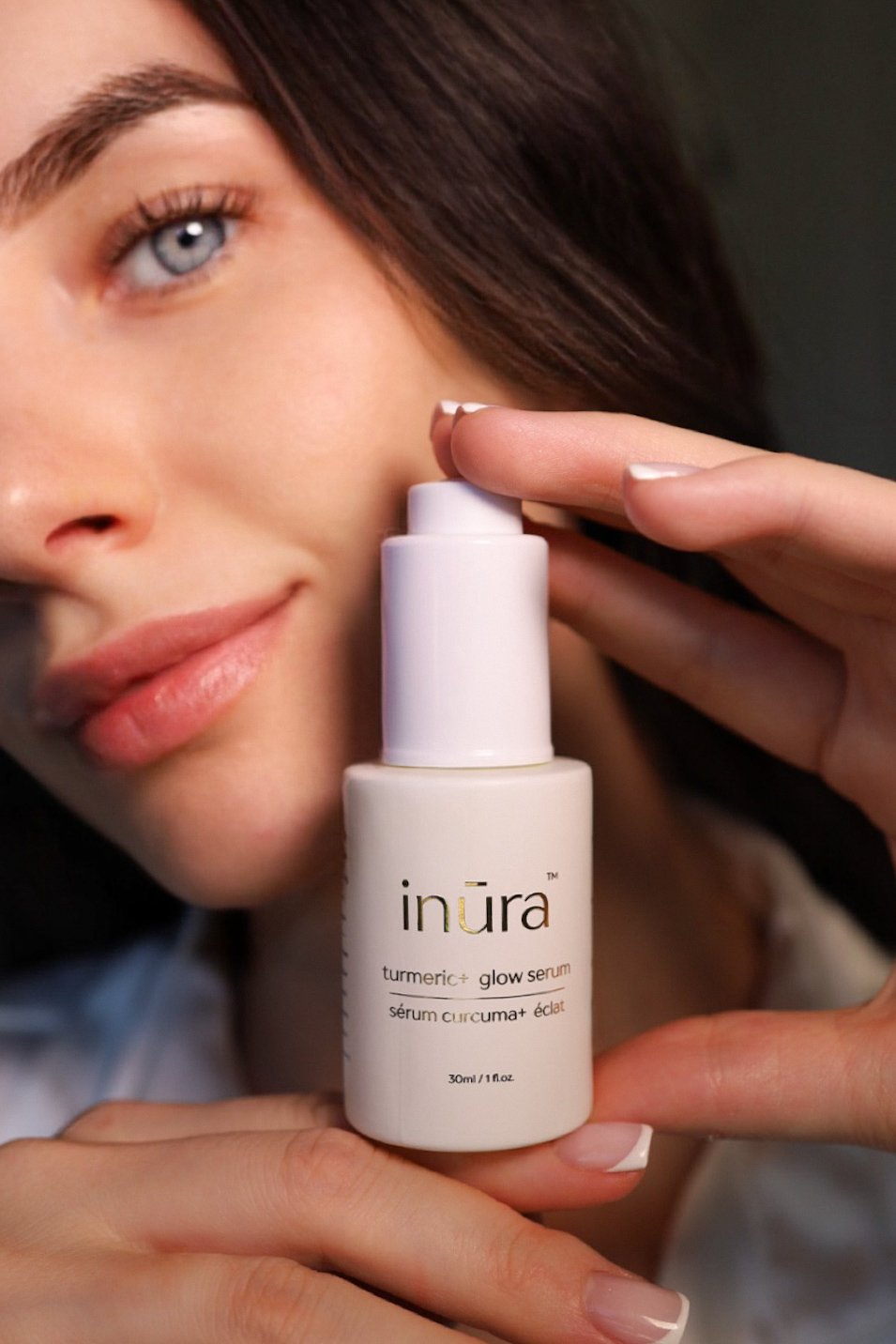 A woman with blue eyes and brown hair holding a white bottle of  inūra turmeric glow serum close to her face.