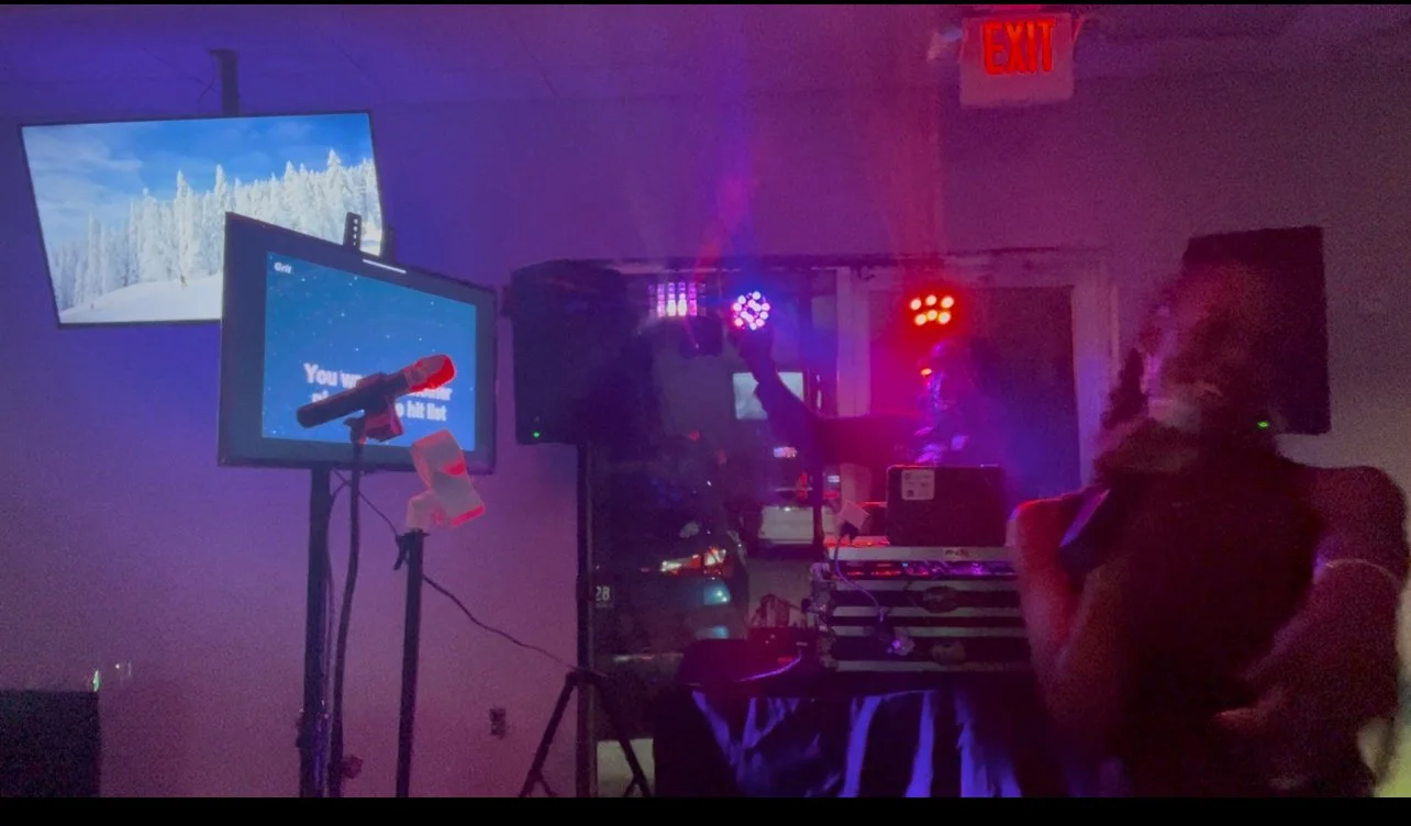 A dimly lit karaoke room with a woman singing into a microphone, a man standing with arms crossed, multiple TVs, colored lights, and a DJ setup.