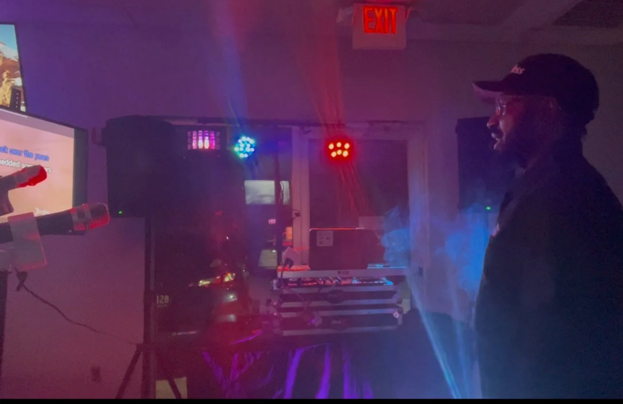 Man in black cap and glasses standing in a room with colorful LED lights, a large speaker, and a screen displaying karaoke lyrics.