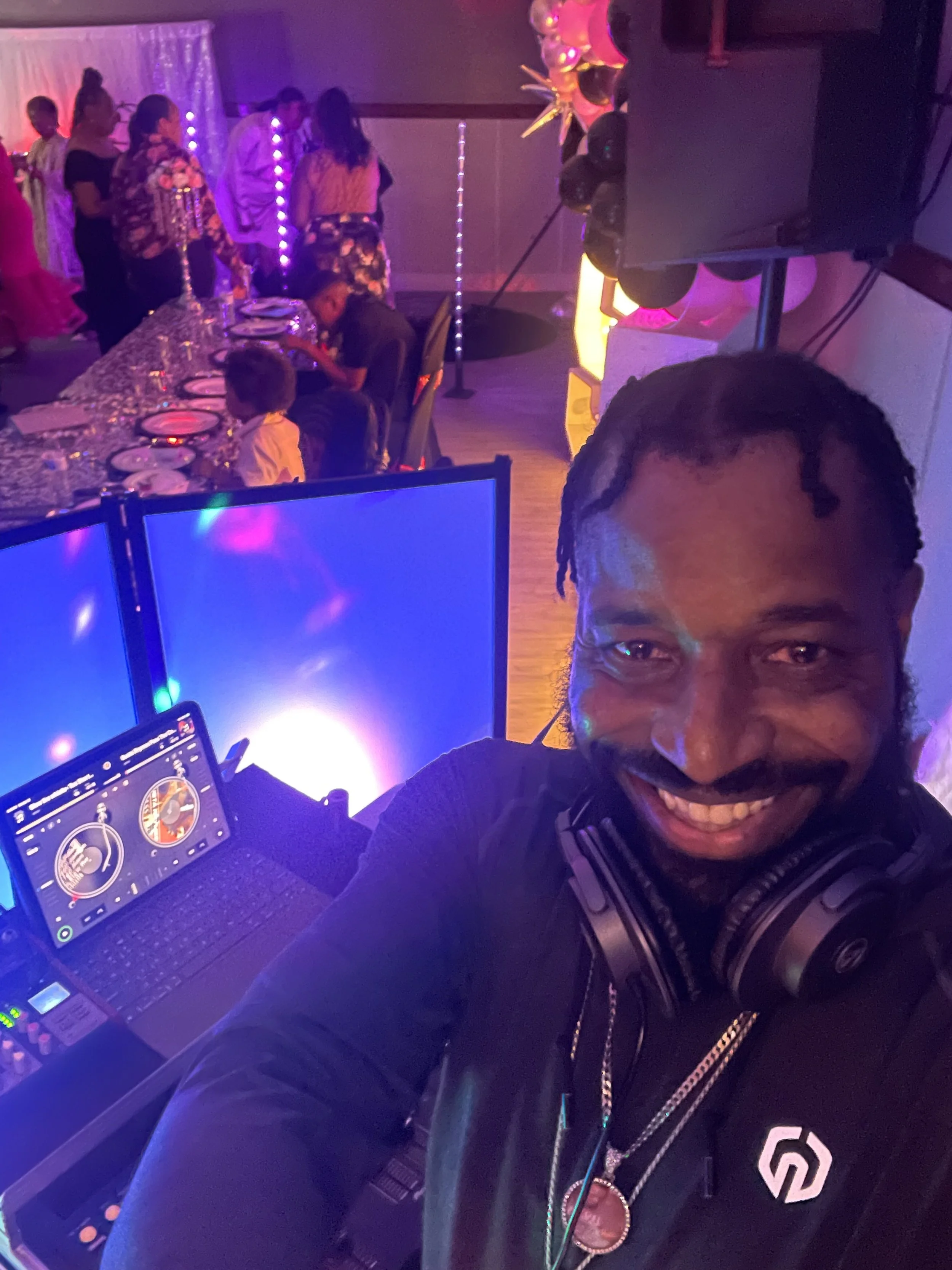 A man wearing headphones is taking a selfie at a party or celebration with children and adults dancing and sitting at a decorated table in the background. The room is lit with colorful, dim lighting, and the man is smiling.