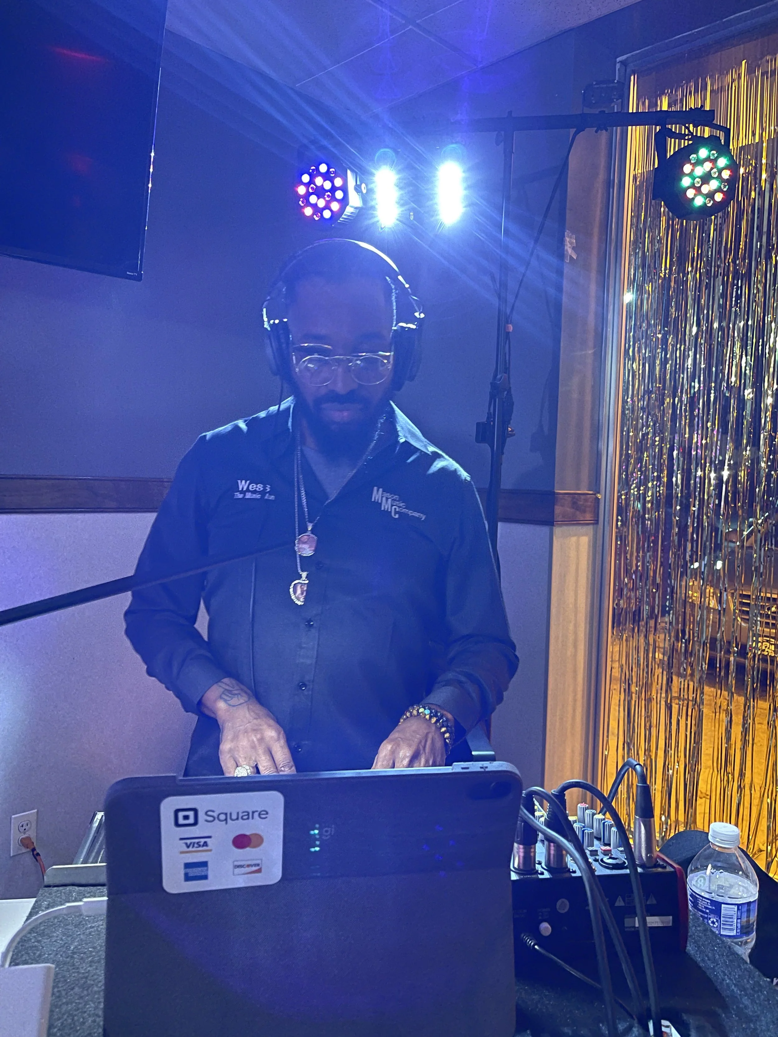 A DJ is standing behind a mixing console with colorful lights in the background, wearing headphones and glasses.