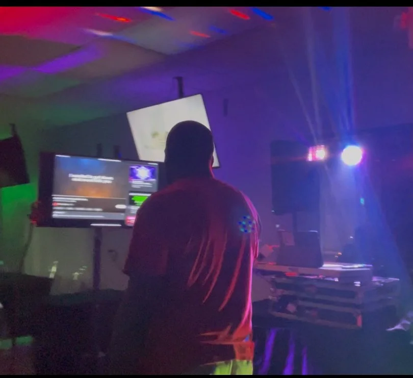 A DJ stands in a dimly lit room with colorful lights, operating DJ equipment and screens, creating a party or club atmosphere.
