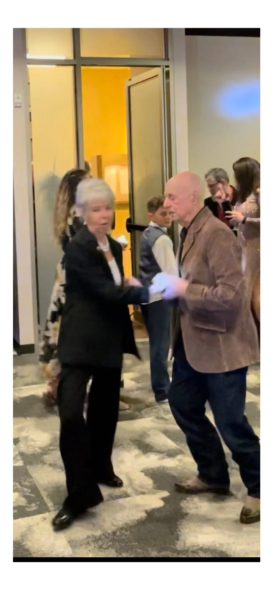 A woman with gray hair and a black suit shaking hands with an older man with a bald head and brown blazer in a crowded indoor event.