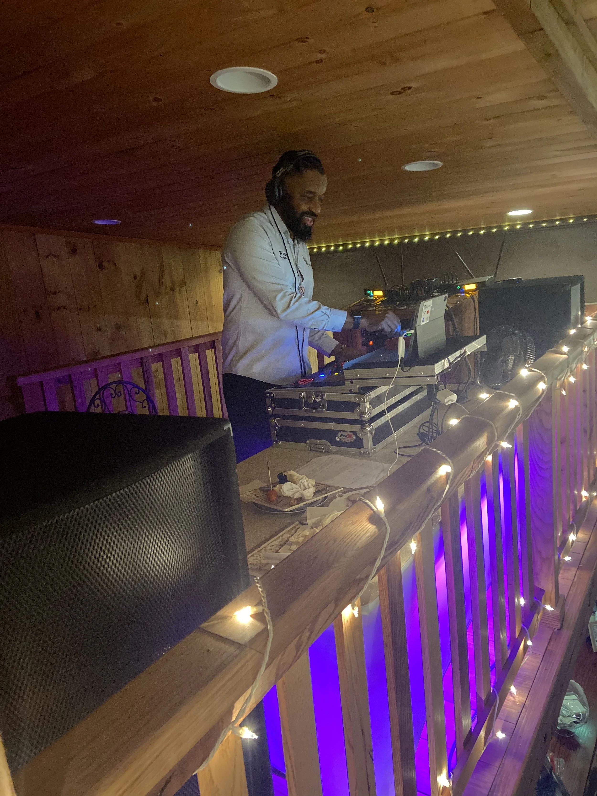 DJ wearing headphones, working with a sound mixer and laptop on a wooden balcony decorated with string lights and purple lighting.