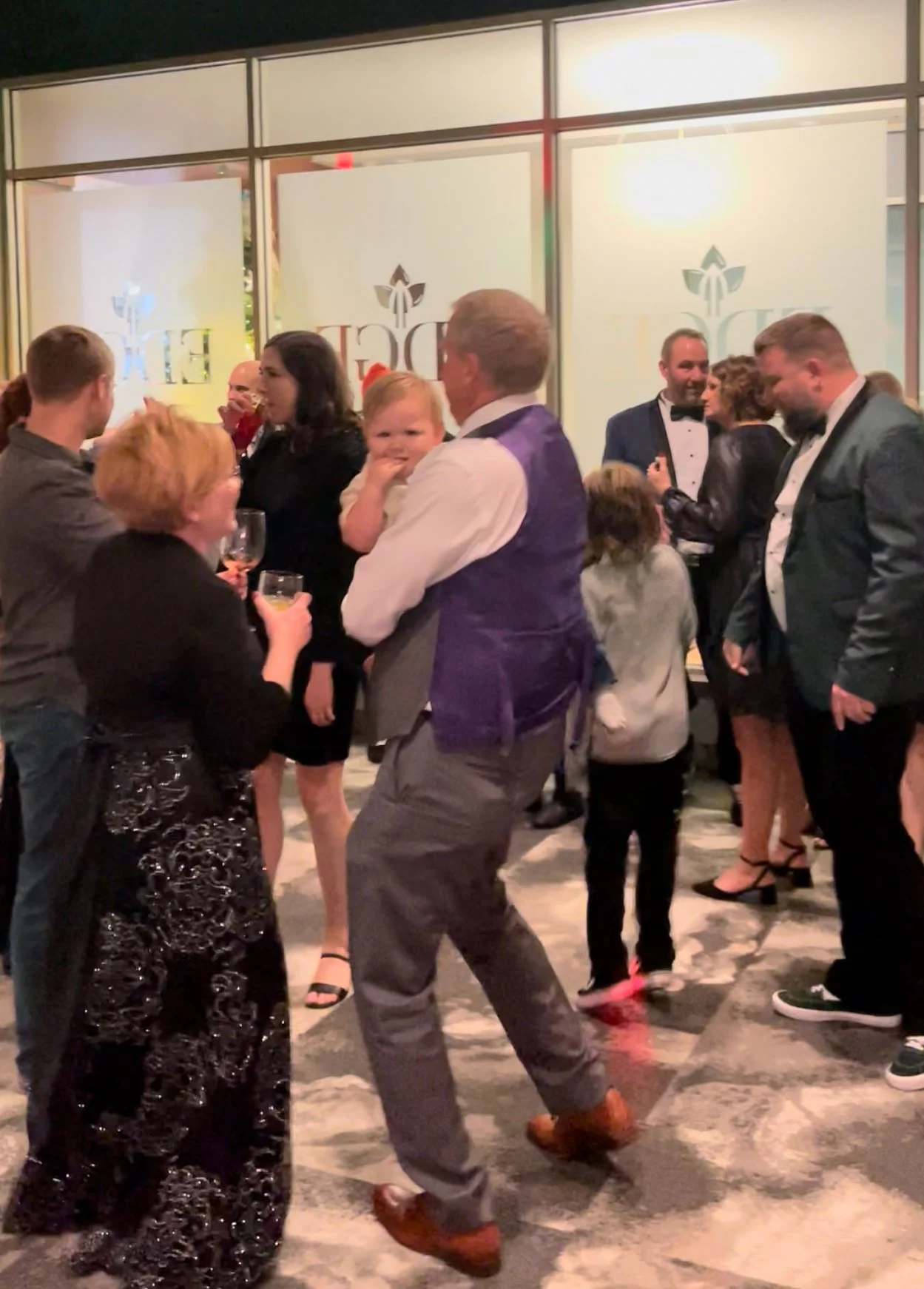 People dancing and socializing at a party or celebration in an indoor venue, with a mix of adults and children, dressed in formal to semi-formal attire.