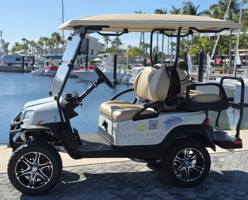An electric golf cart parked near a marina with boats, water, palm trees, and buildings in the background. The cart has beige seats, a roof, and branding for Oceans Edge Resort & Marina.