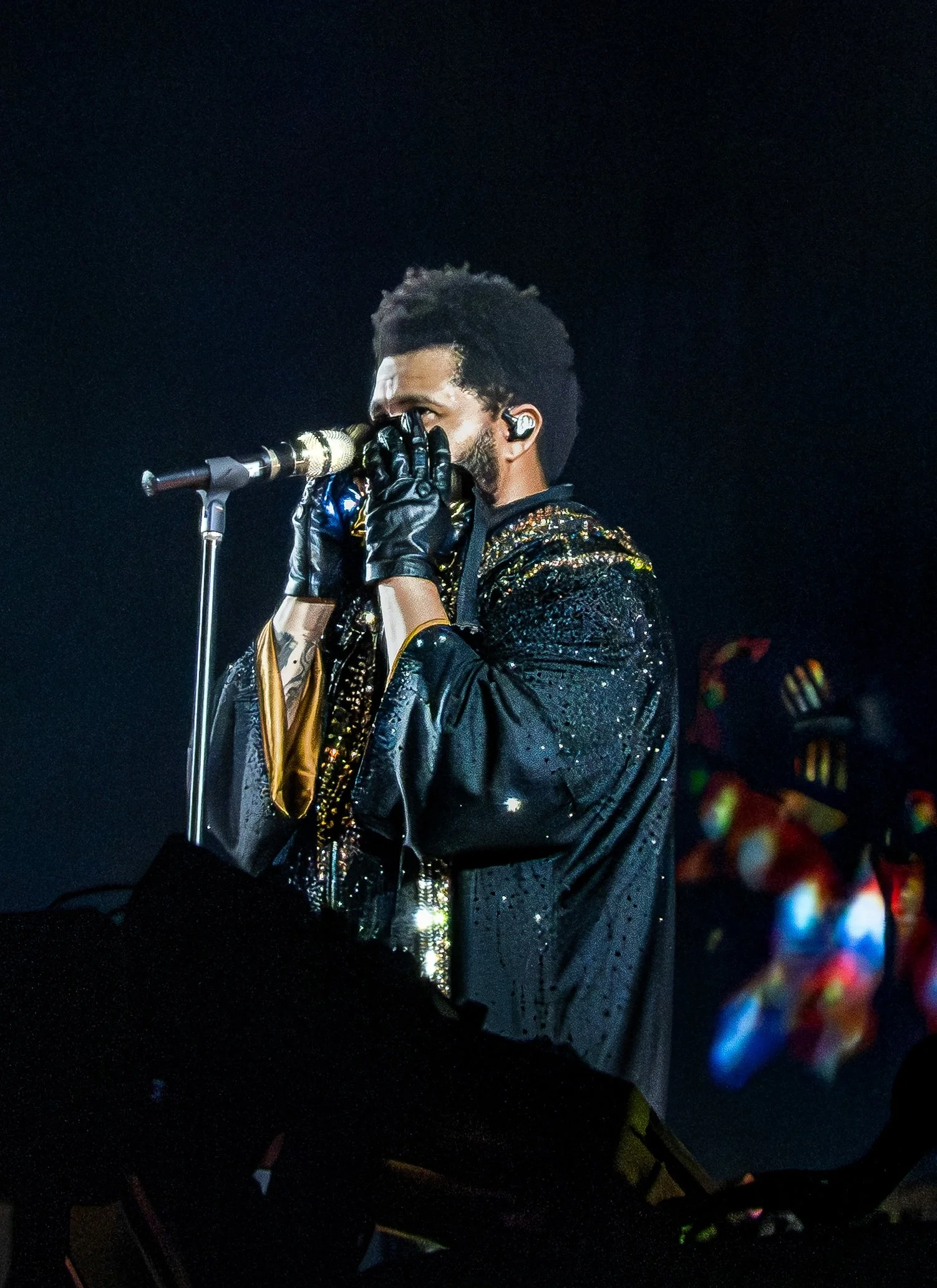Concert portrait of a singer, The Weeknd, performing into a microphone under dramatic stage lighting.
