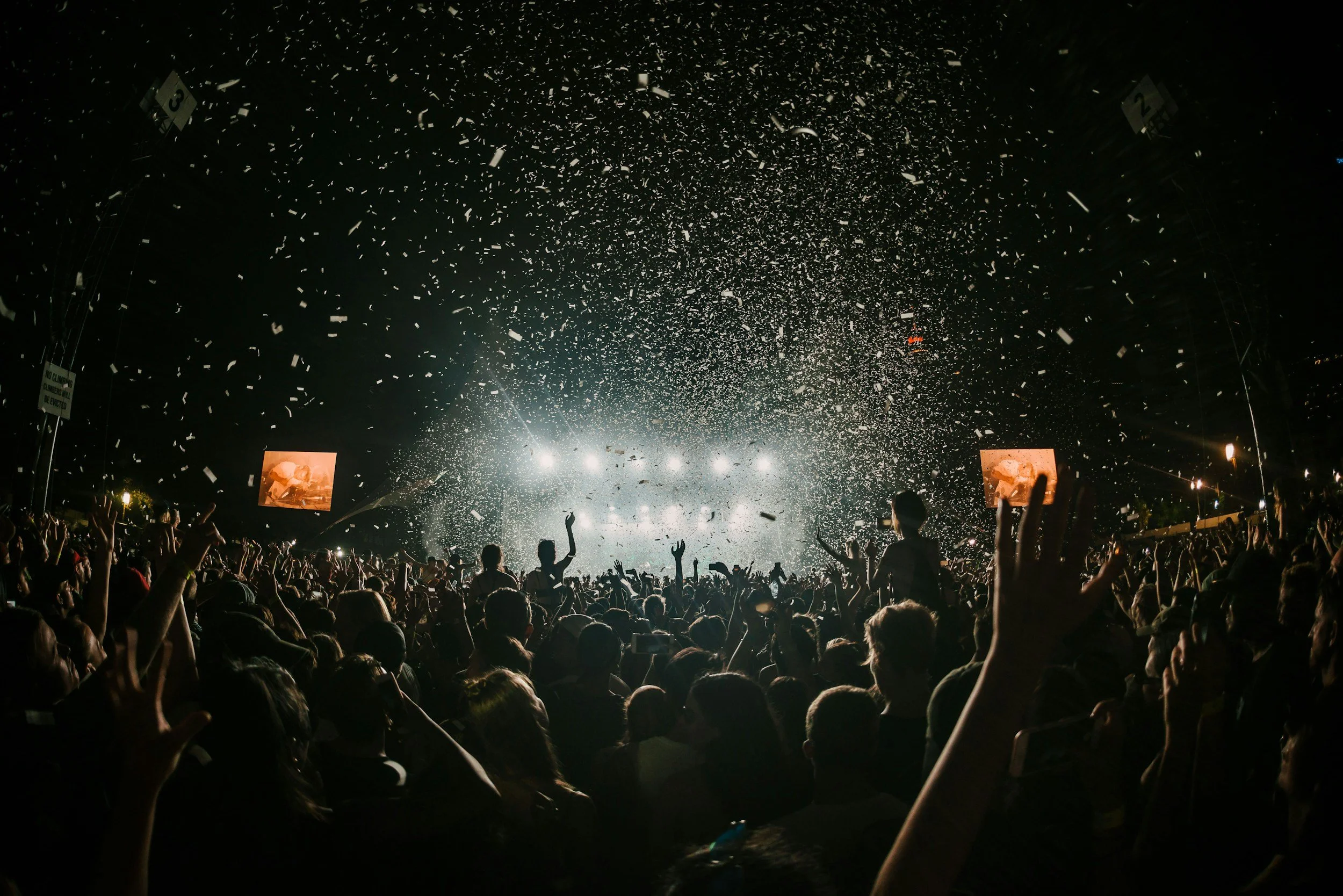 Crowd at a concert or festival at night with confetti falling and bright lights behind the stage.
