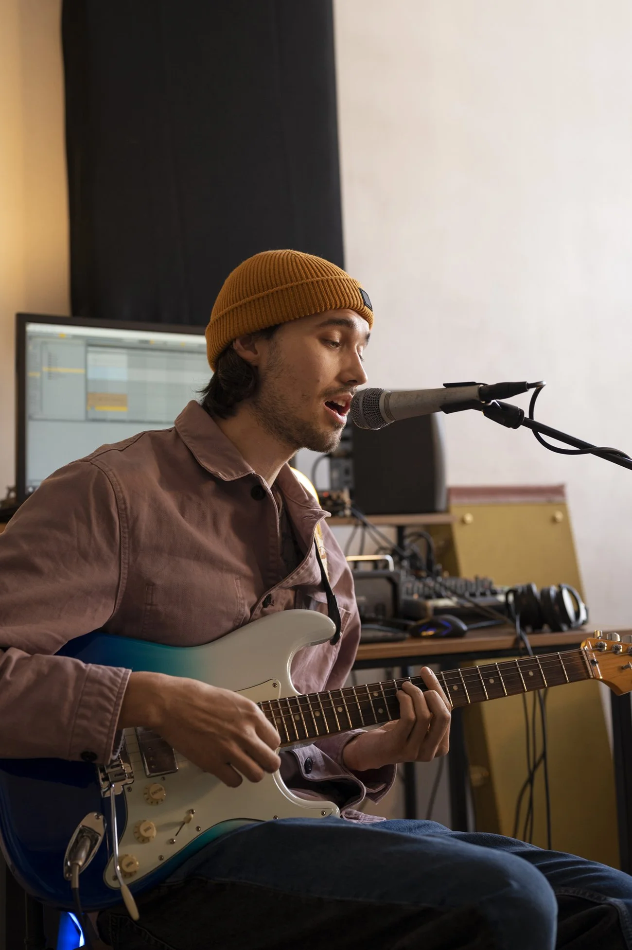A man wearing a mustard yellow beanie and a pinkish-brown shirt is playing an electric guitar and singing into a microphone in a music studio.