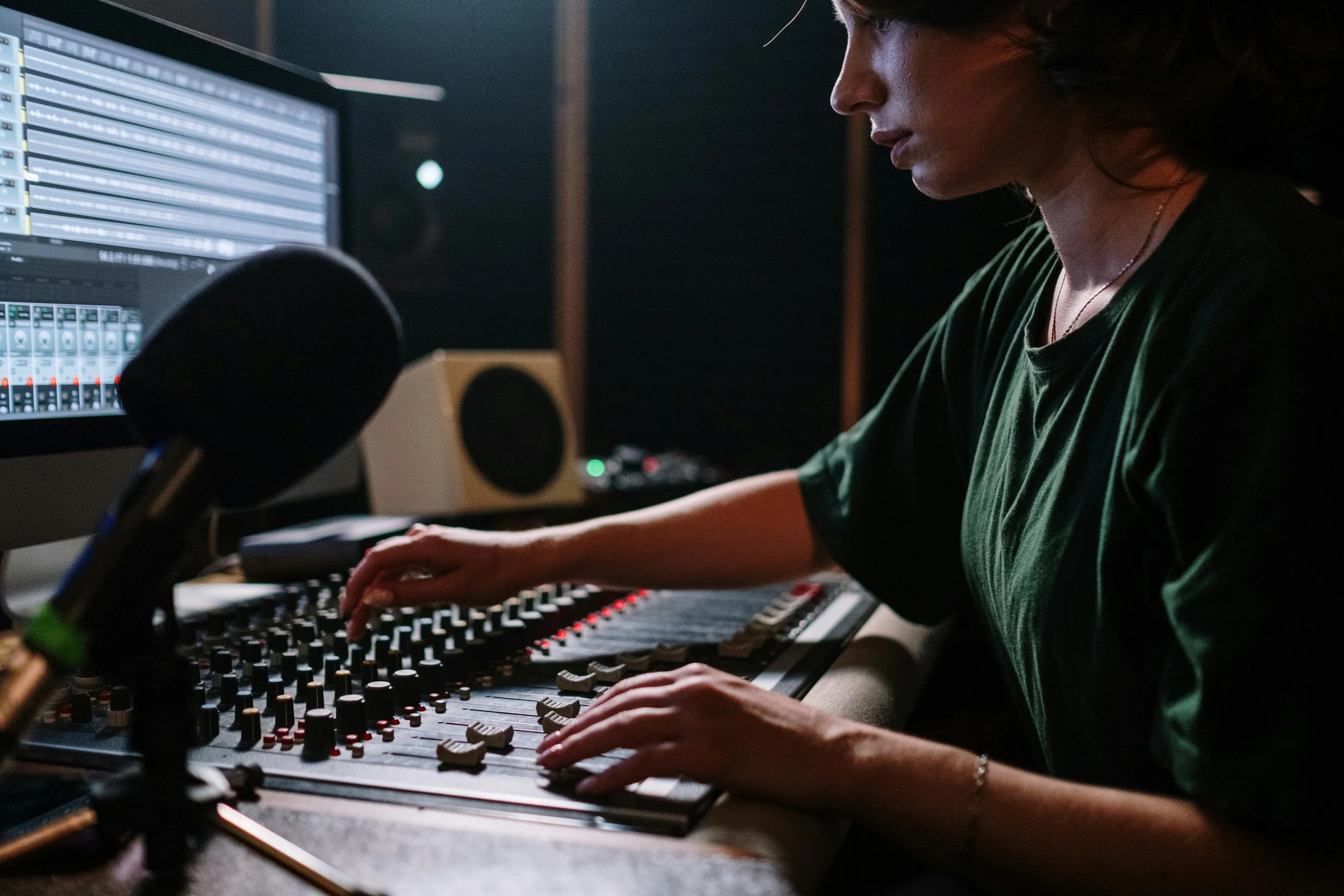 A person wearing a green shirt working with audio mixing equipment in a recording studio, with a microphone in foreground and computer screens in the background.