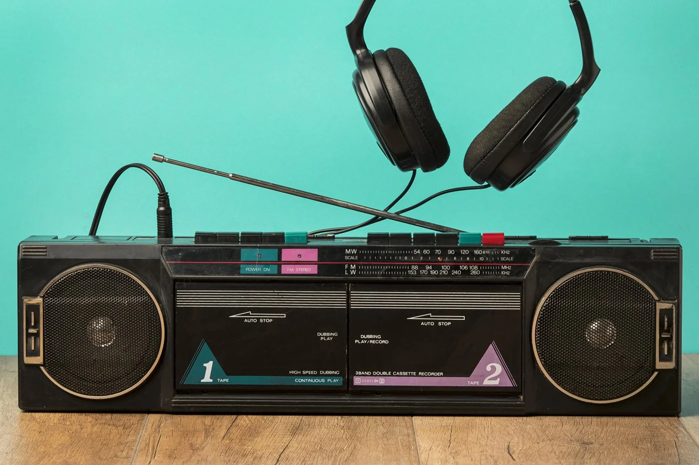 A vintage portable boombox with two speakers, a cassette player, and a pair of black over-ear headphones resting on top, placed on a wooden surface against a teal background.