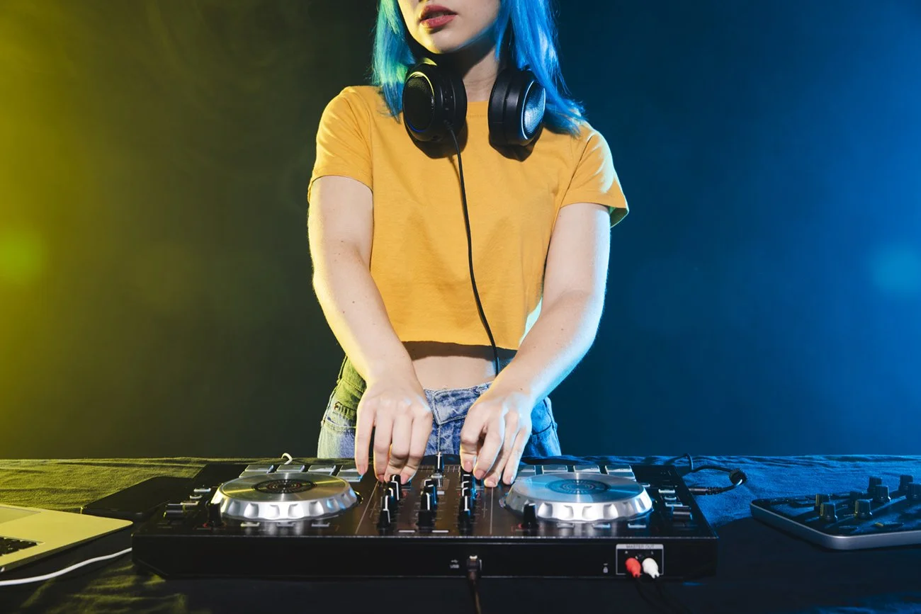 Person with blue hair wearing an orange shirt and headphones, DJing with a mixer and laptop at a colorful party or club.