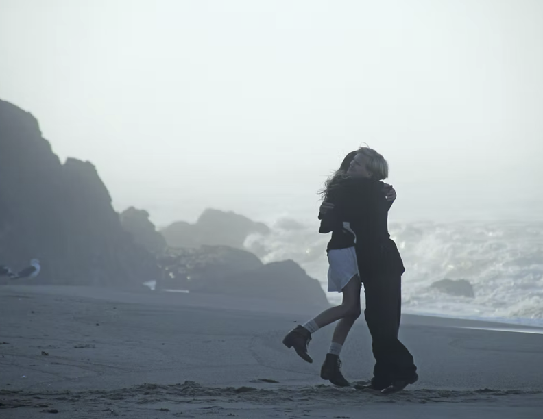 A couple hugging on a foggy beach with rocks and the ocean in the background.