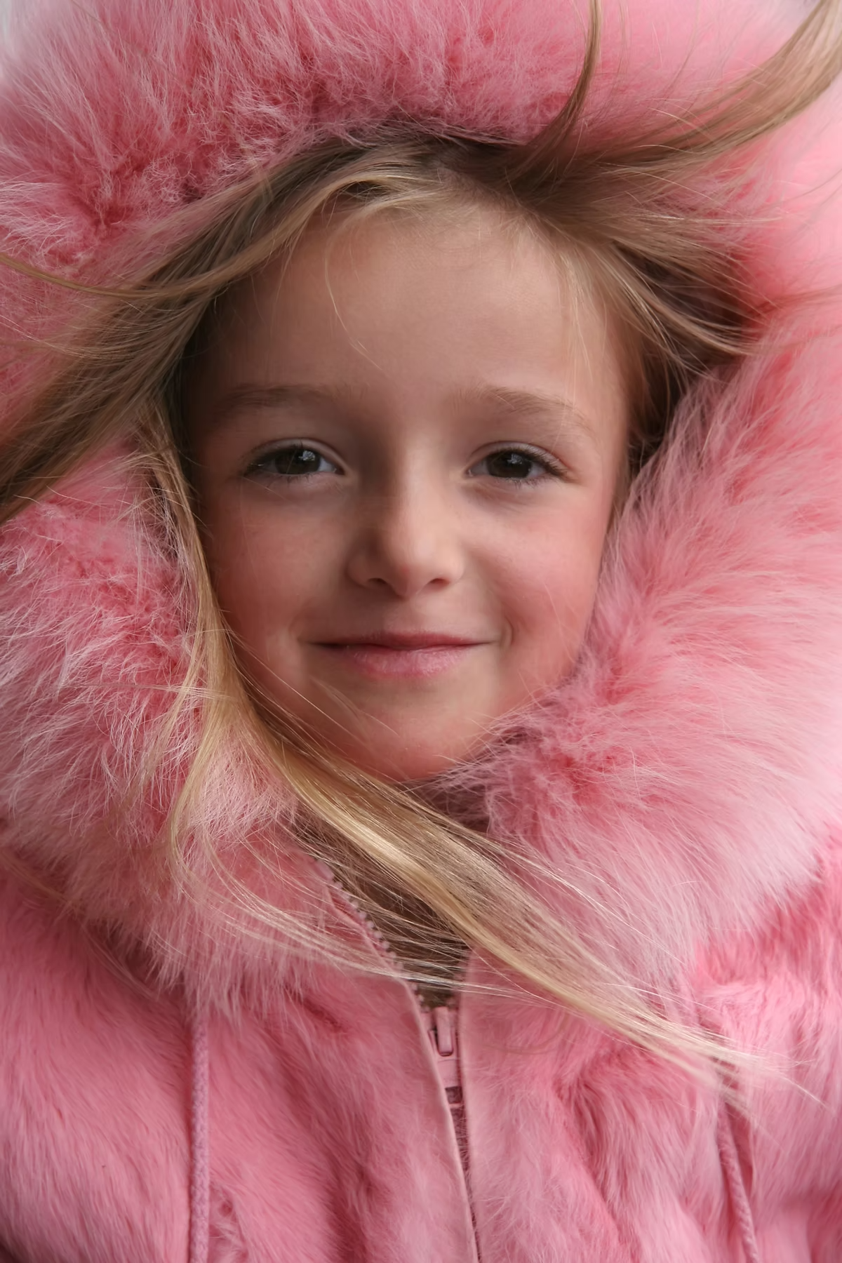 A young girl with blonde hair smiling, wearing a pink fluffy coat with the hood up.