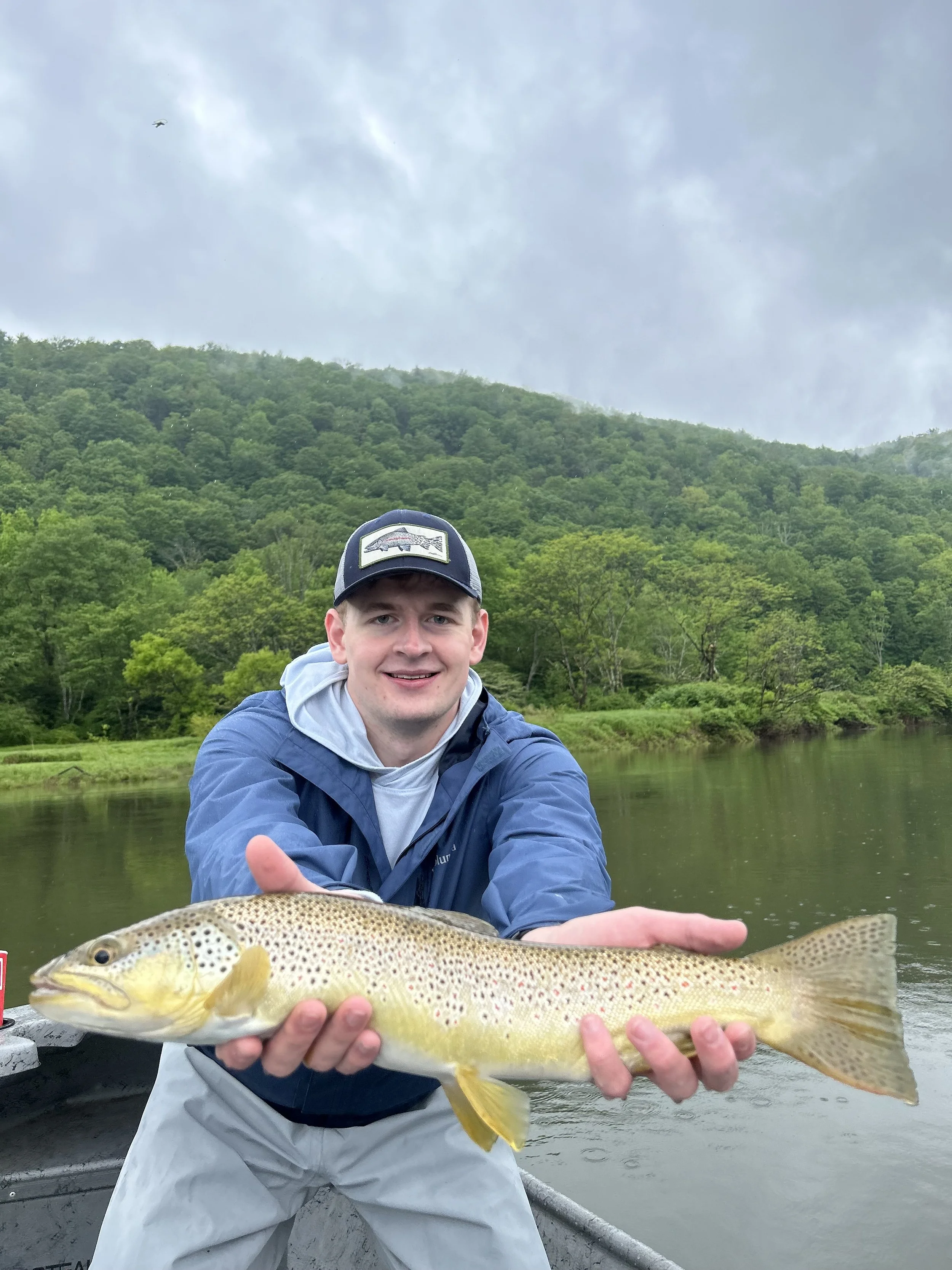 Young man smiling while holding a large trout fish on a boat with a river and green forested hills in the background.