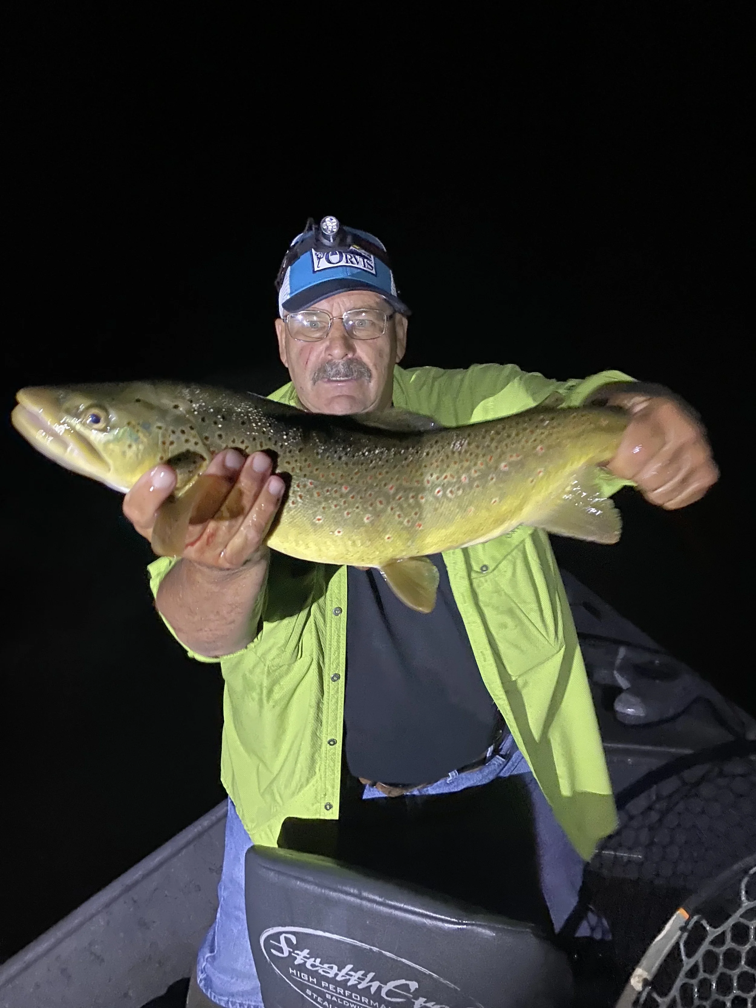 Man in a headlamp and bright yellow jacket holding a large brown trout at night.