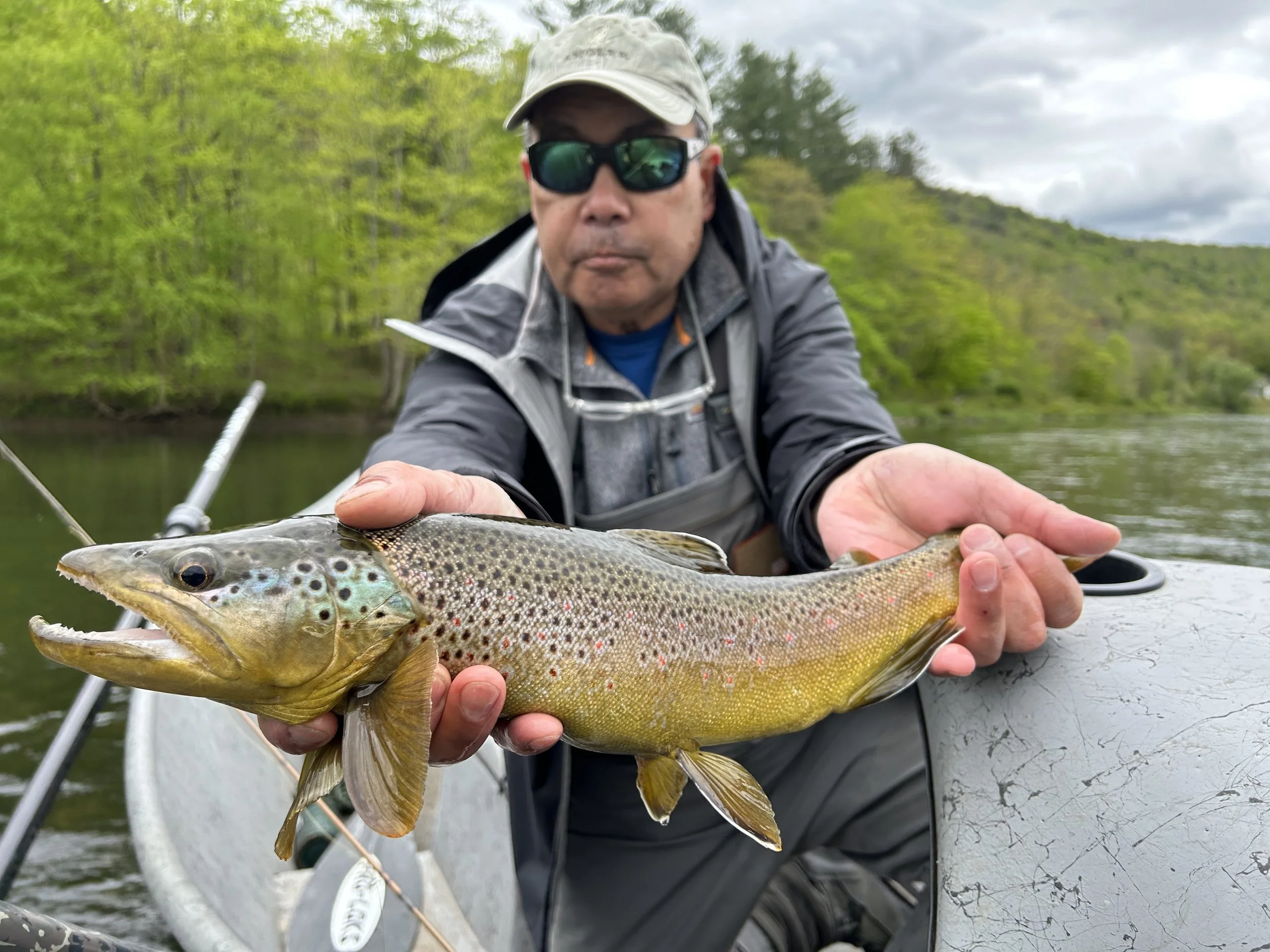 A man wearing sunglasses, a gray jacket, and a beige cap holding a caught fish on a boat with a background of a river and green trees.