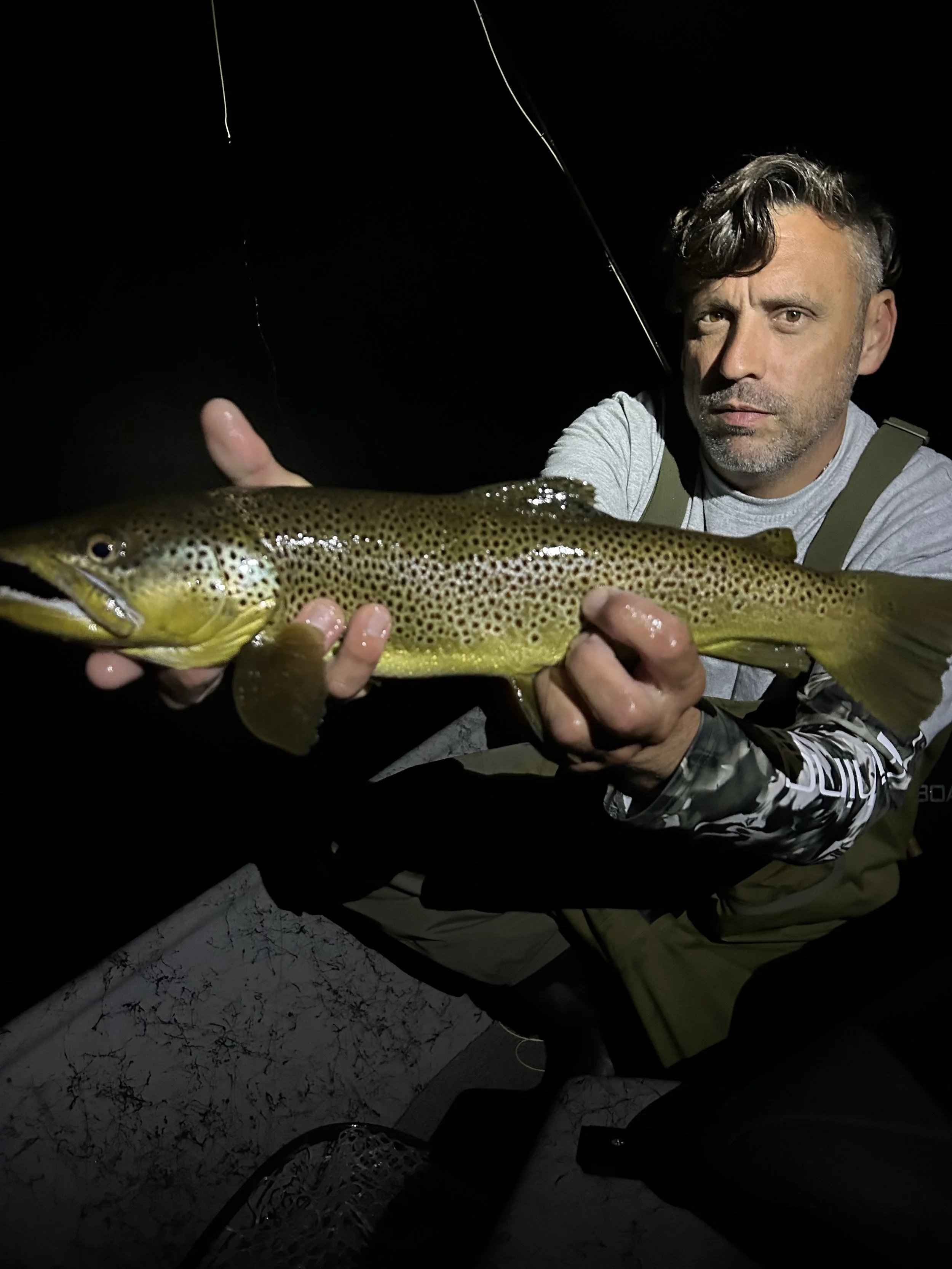 Man holding a large fish at night, with a fishing rod above him, on a boat with a marbled surface.