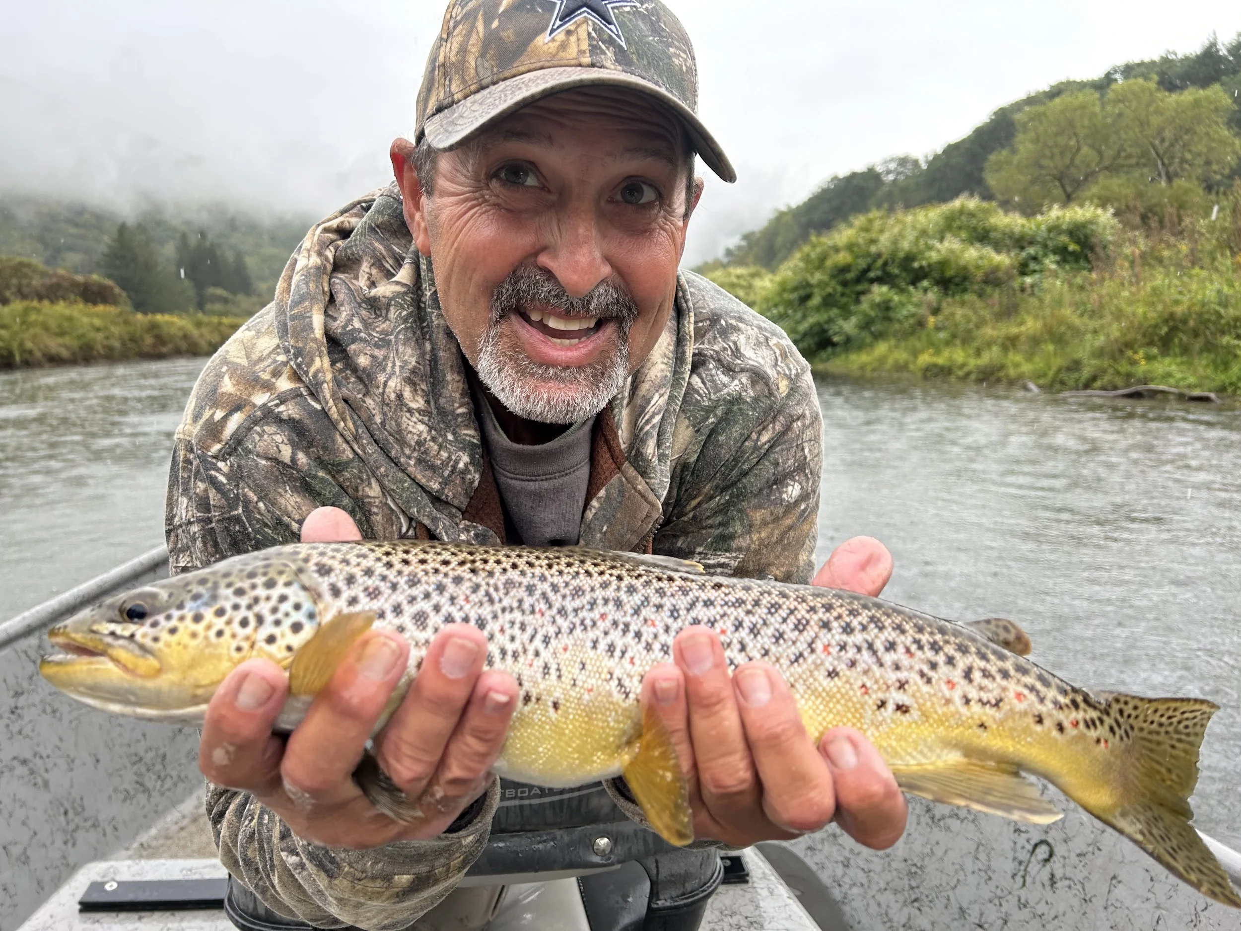 A man in camouflage clothing holding a large rainbow trout fish on a boat, with a river and green landscape in the background.