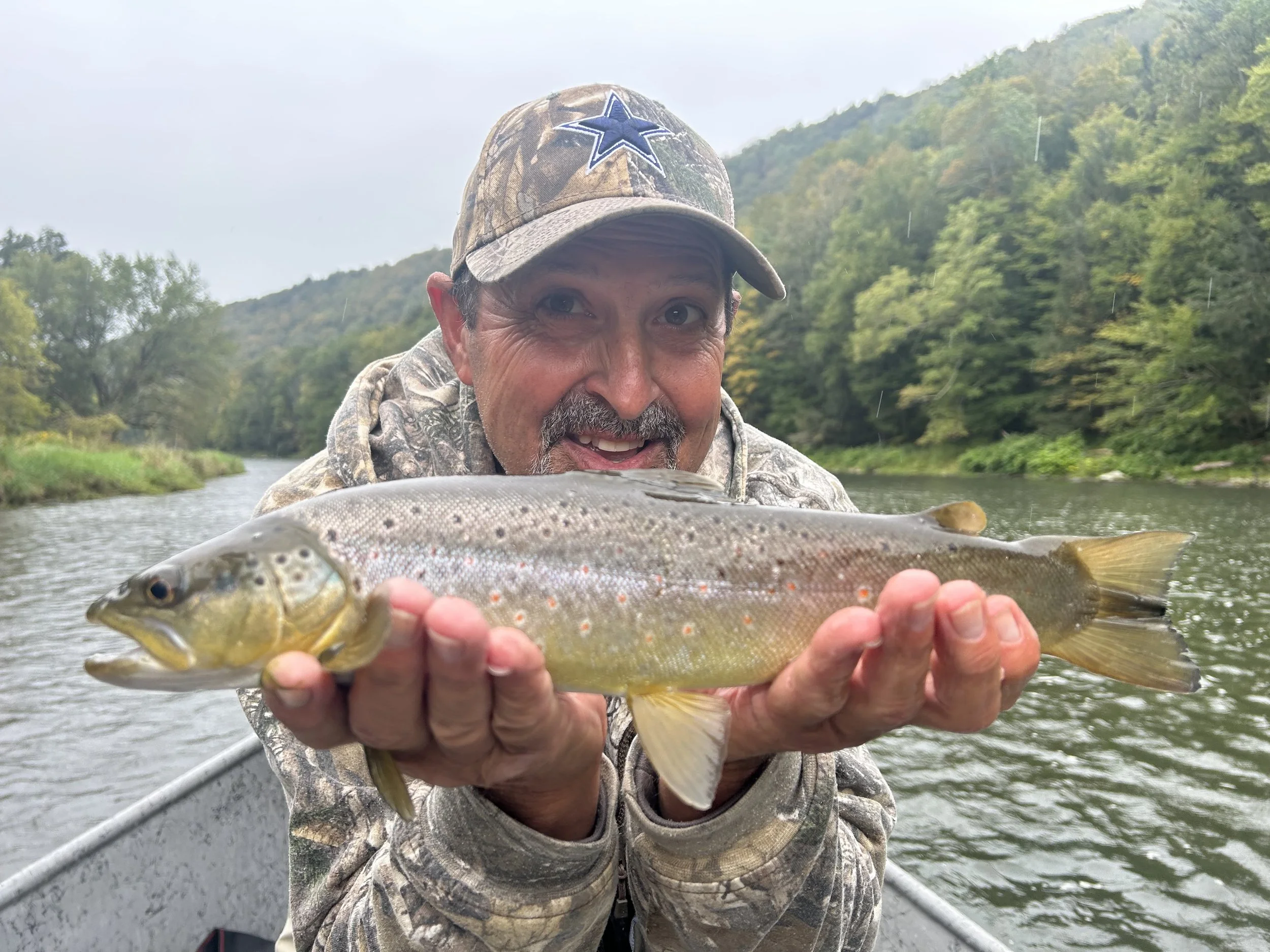 A man in camouflage clothing and a camouflage cap holding a fish with green and brown coloring while in a boat on a river, with green trees and hills in the background.