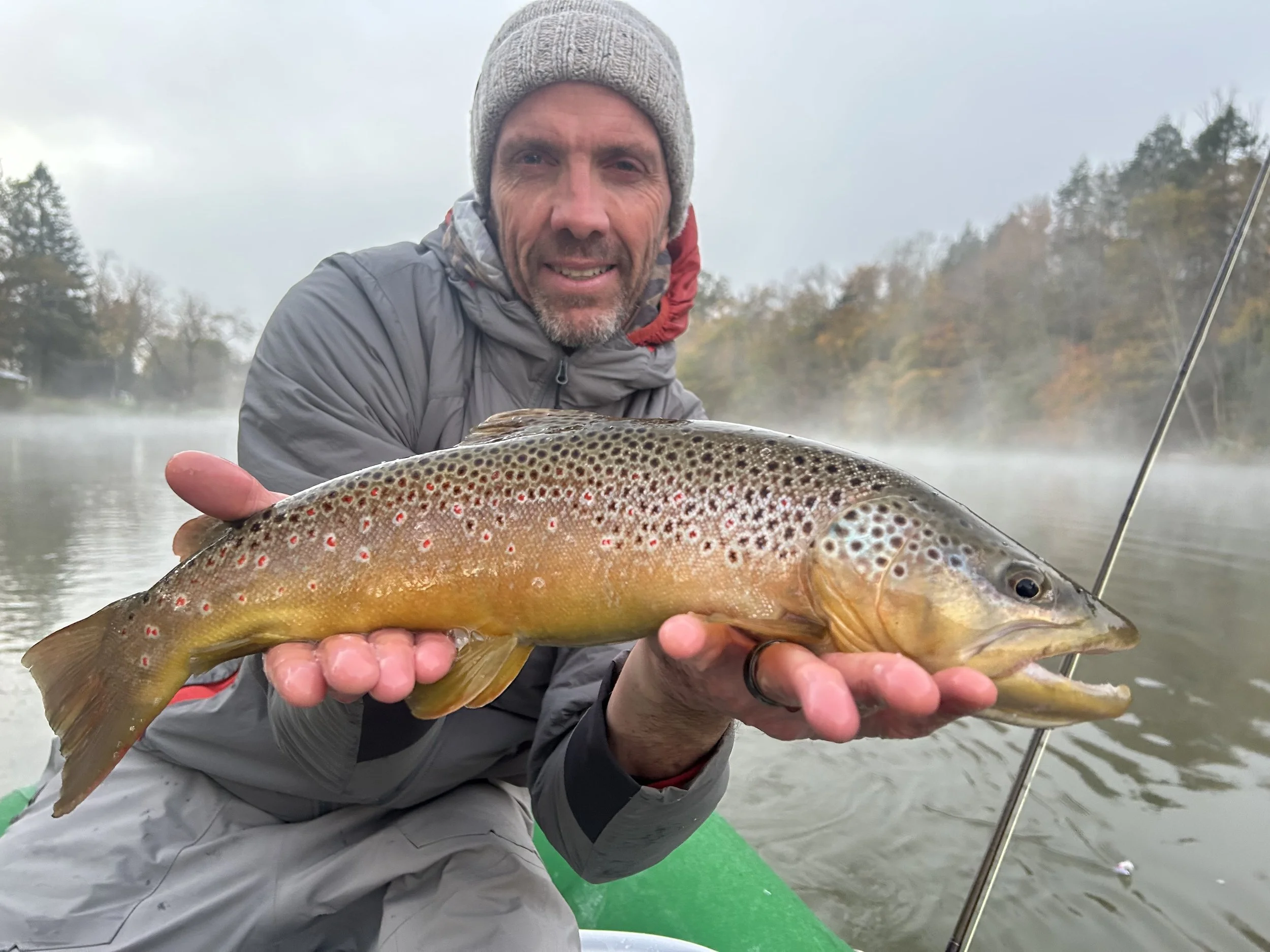 Man in gray jacket and beanie hat holding a large brown and yellow trout fish while on a boat in a misty river with trees in the background.