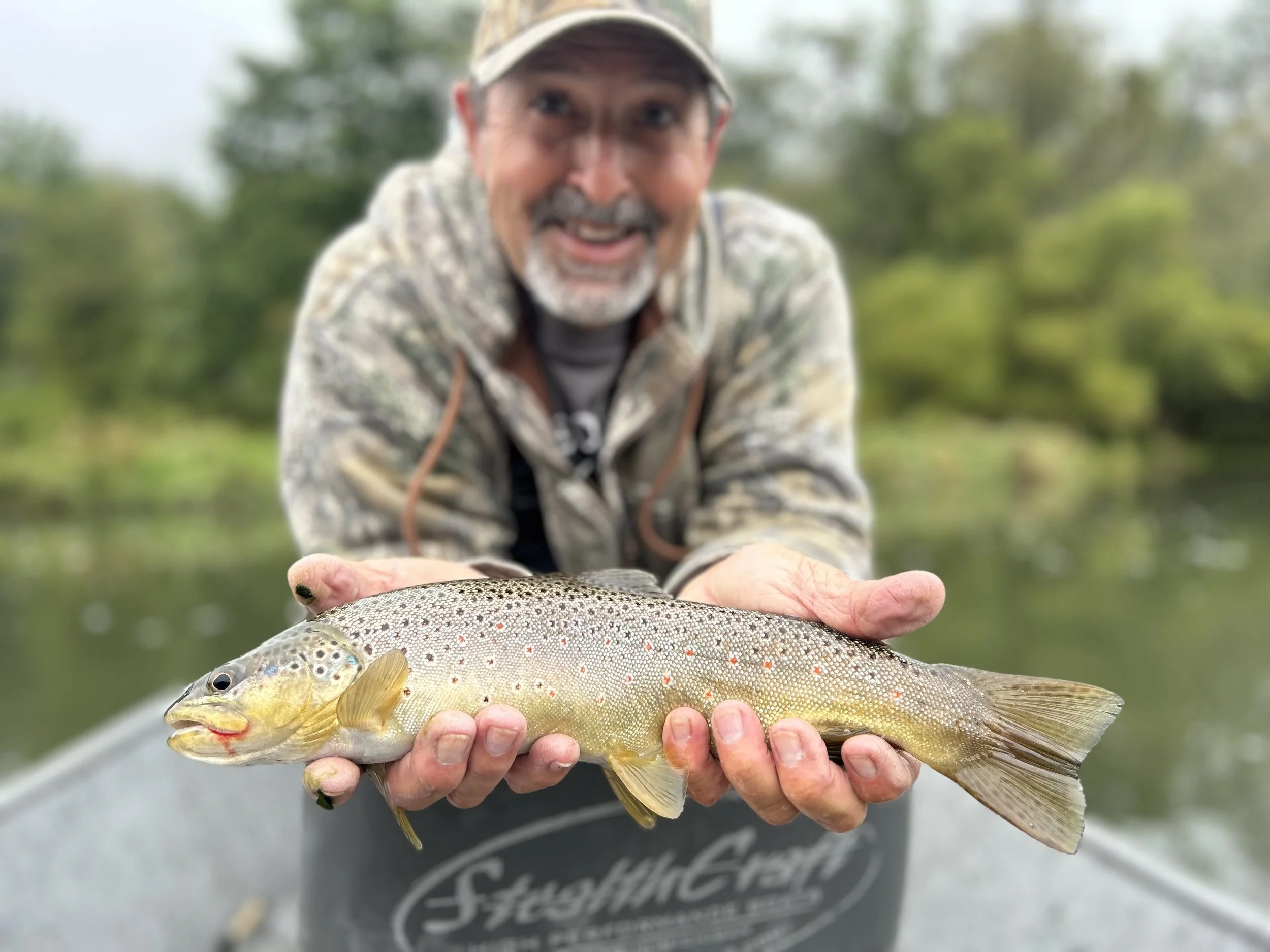 Man in camouflage jacket holding a freshly caught rainbow trout fish on a boat with a blurred background of trees and water.
