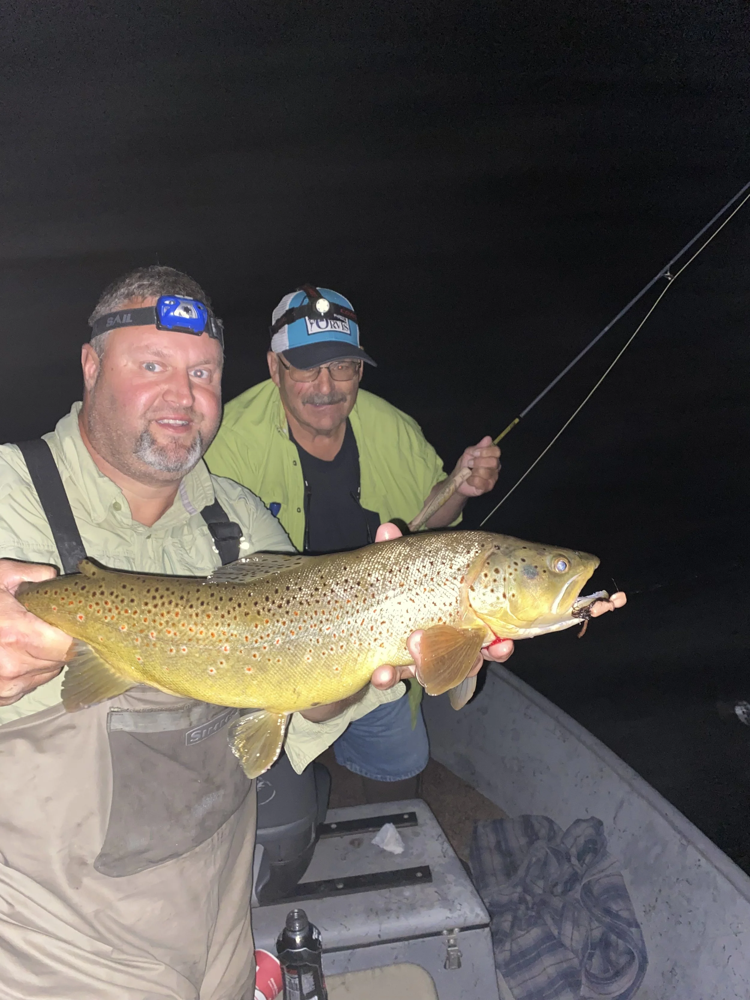 Two men on a boat at night holding a large brown trout with orange spots, one man in a yellow jacket and headlamp, the other in a blue cap with headlamp, using a fishing rod.