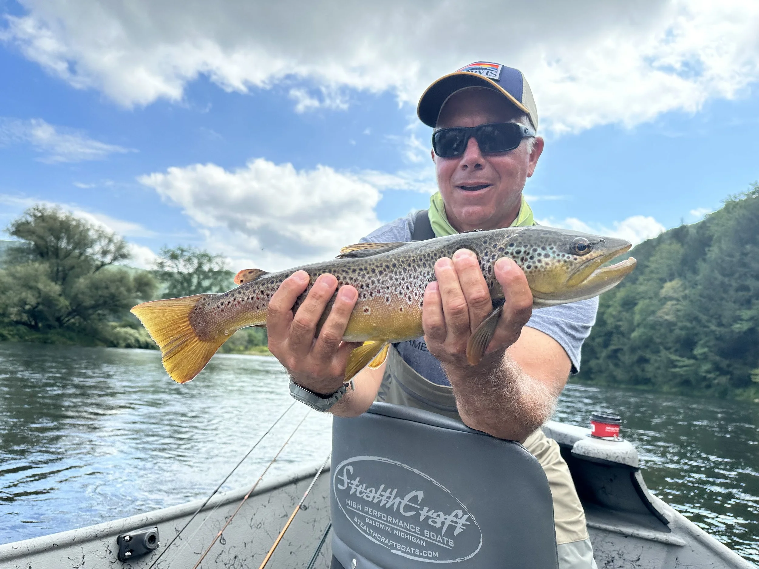 Man wearing sunglasses and a cap holding a fish on a boat in a river with trees and hills in the background.