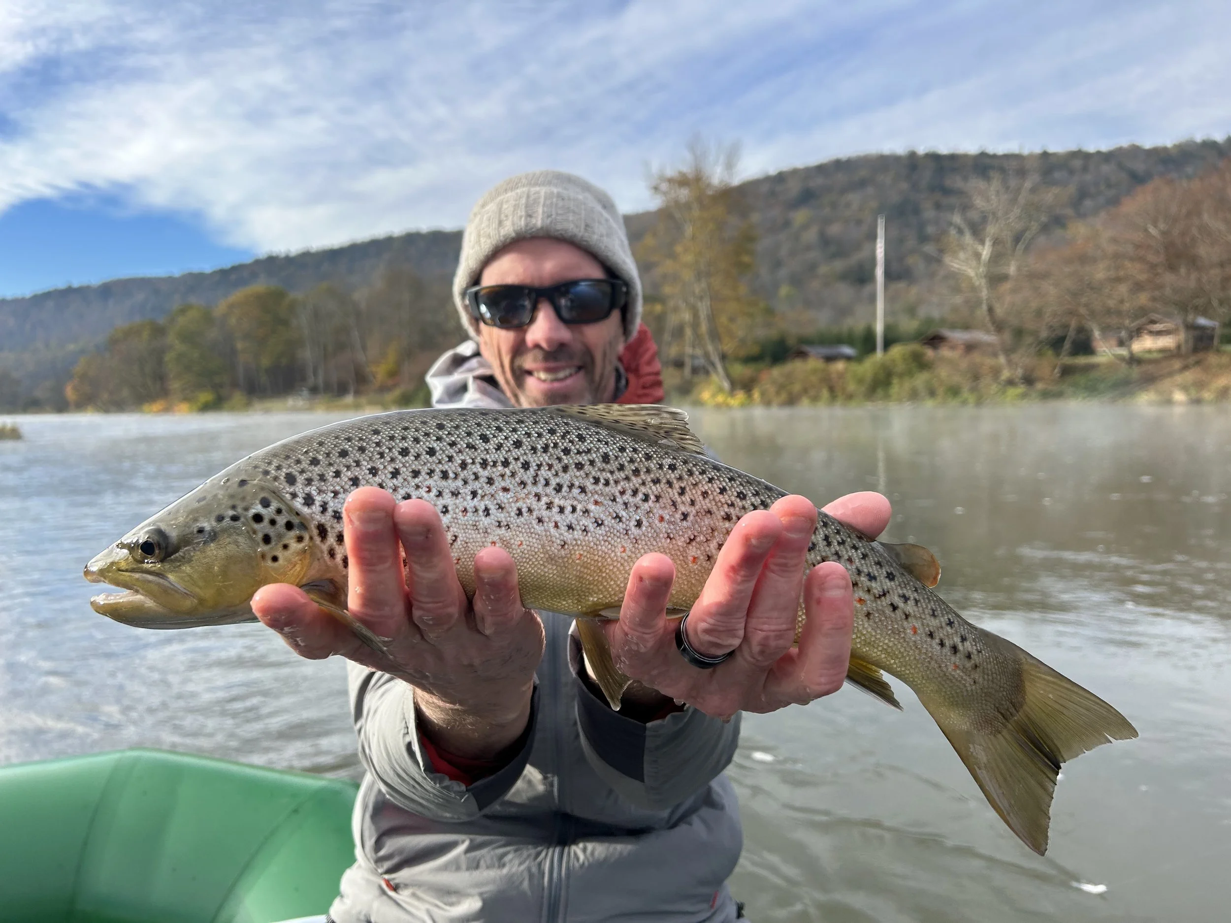 Man wearing sunglasses and a beanie hat holding a large trout fish on a river with hills and trees in the background.