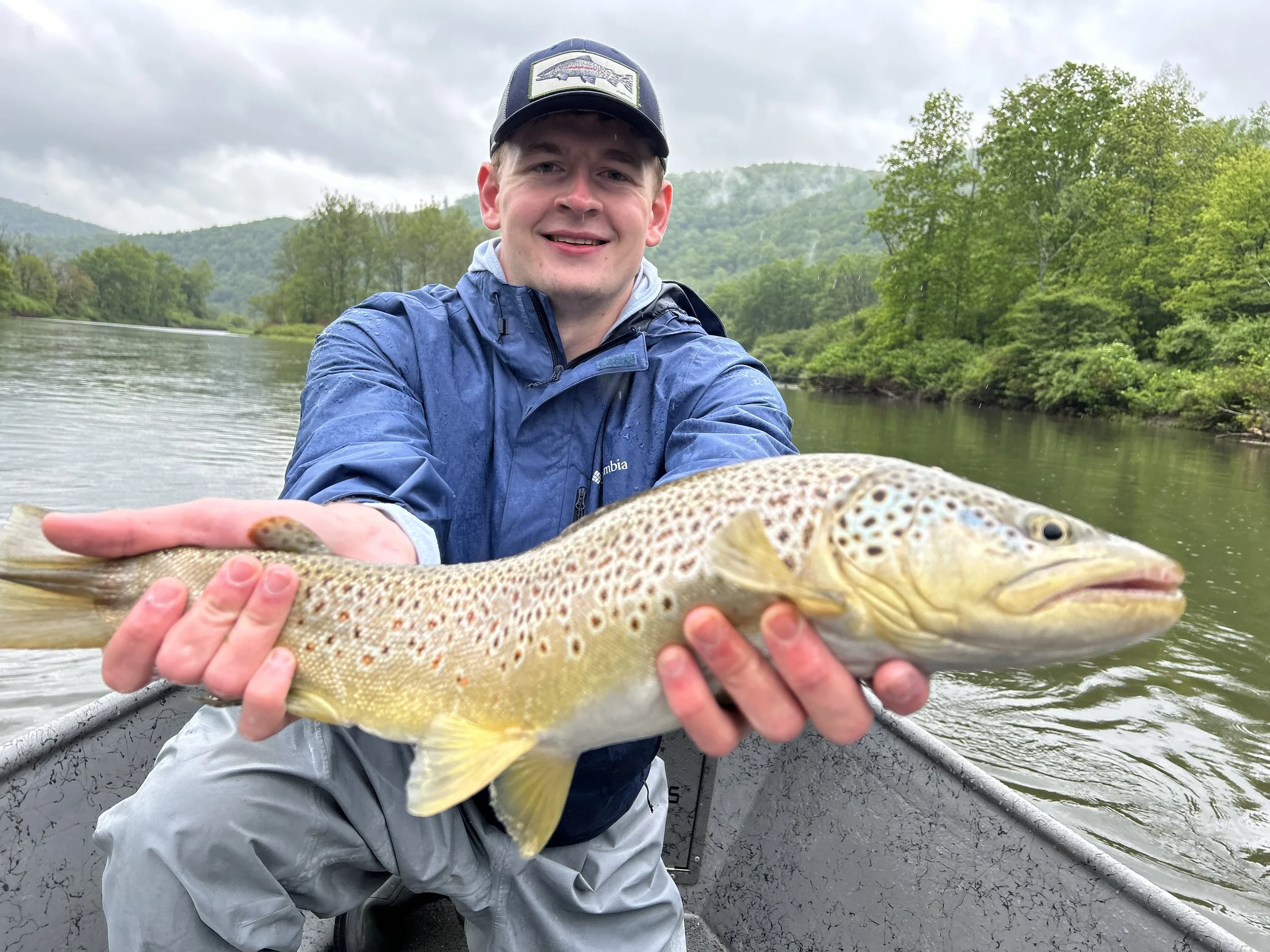 A man in a blue jacket and cap on a boat holding a large spotted fish, with a river and green trees in the background.