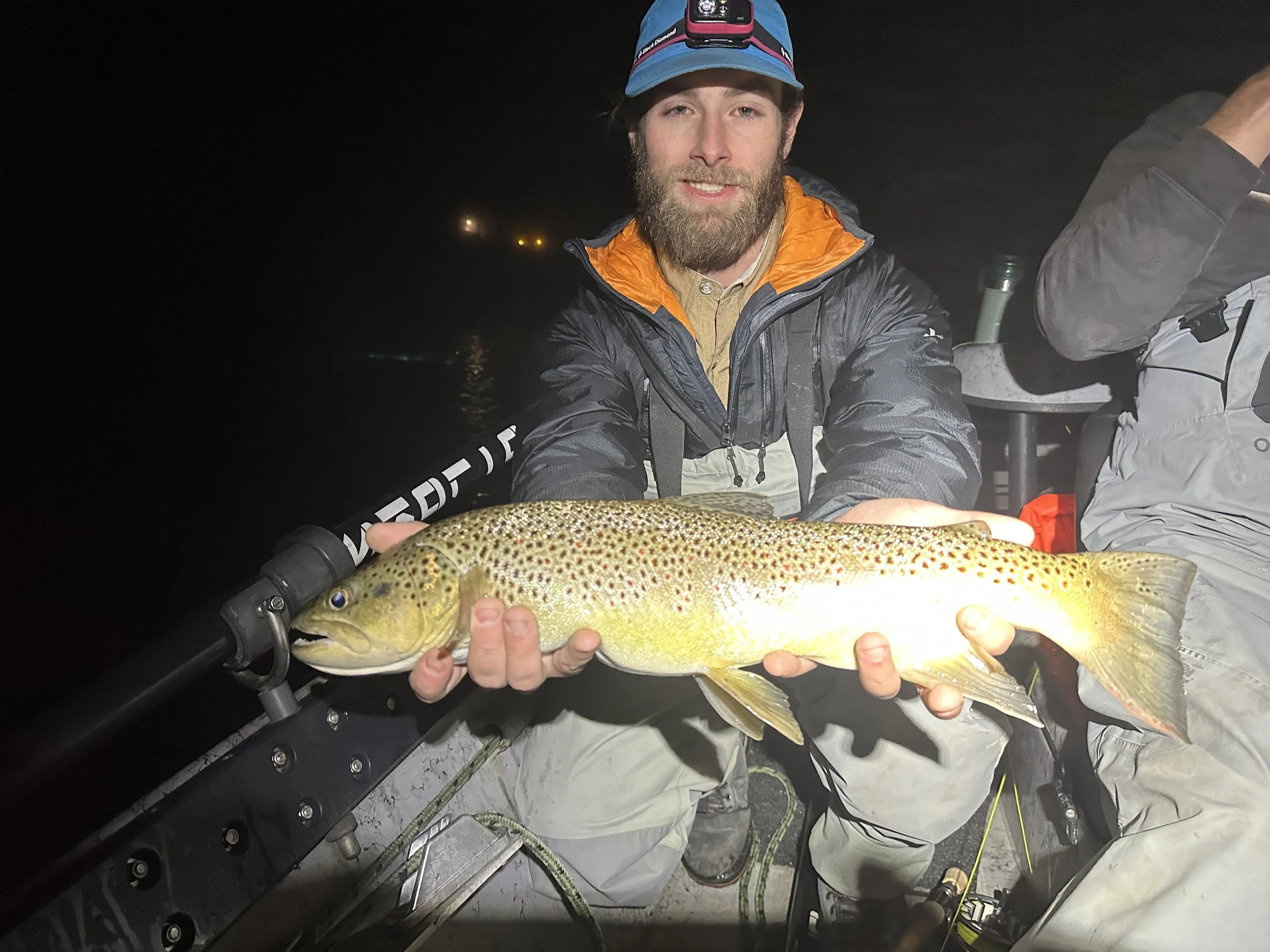 A man wearing a blue cap and black jacket holding a large rainbow trout on a boat at night.