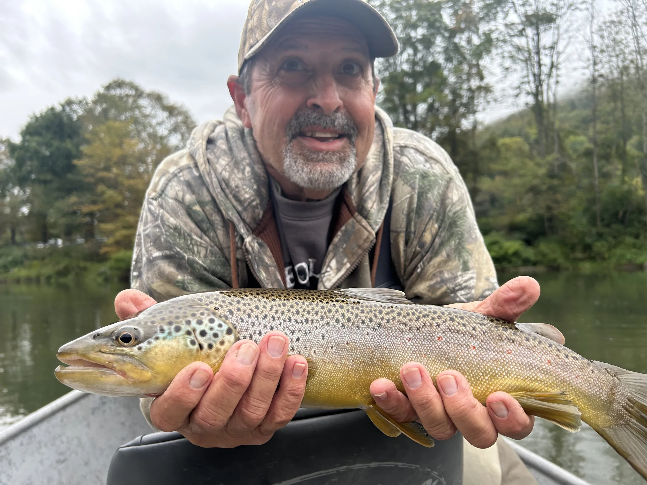 A man wearing camouflage clothing holding a large rainbow trout fish on a boat with a river and trees in the background.