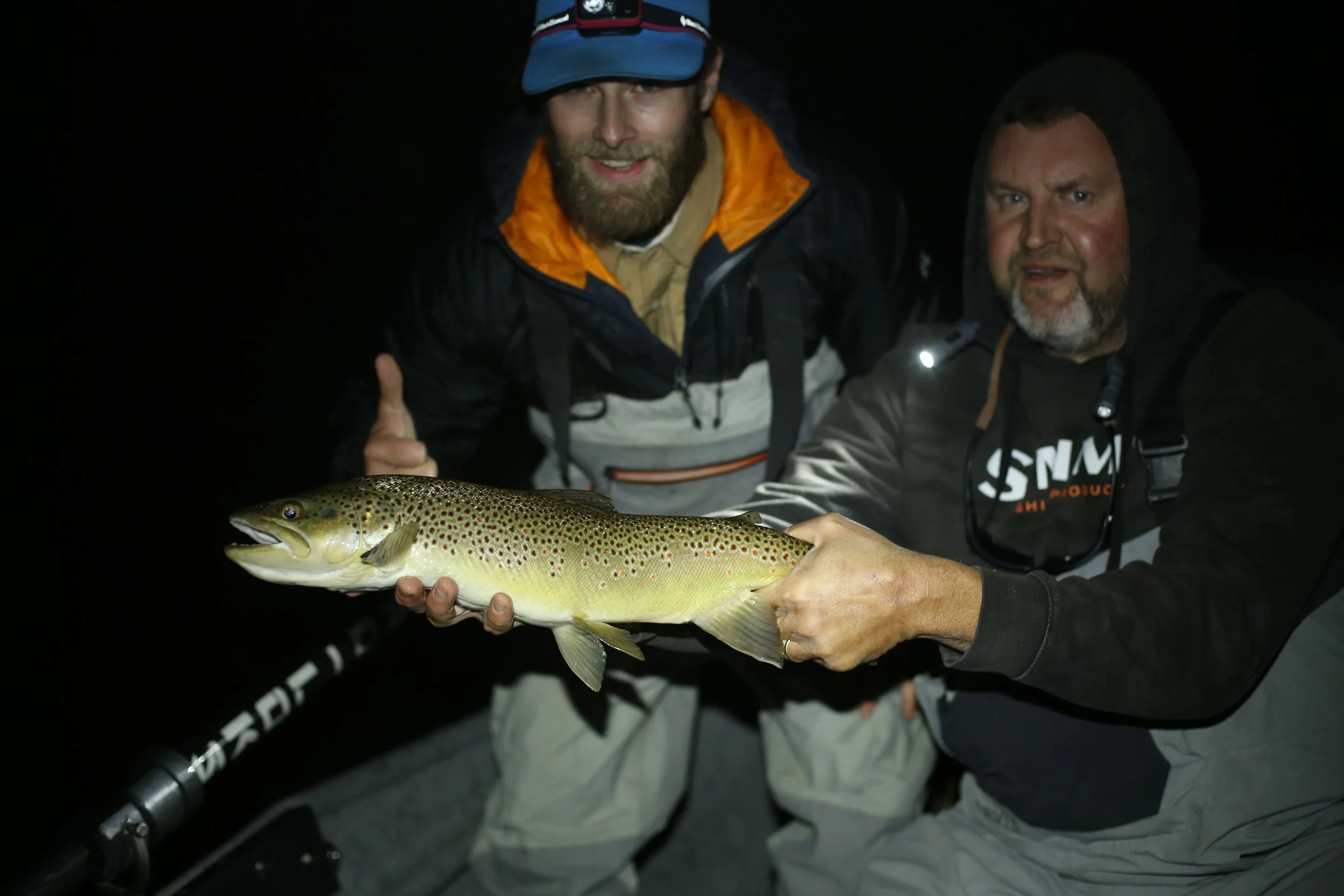 Two men holding a large fish at night during a fishing trip, with one giving a thumbs-up.