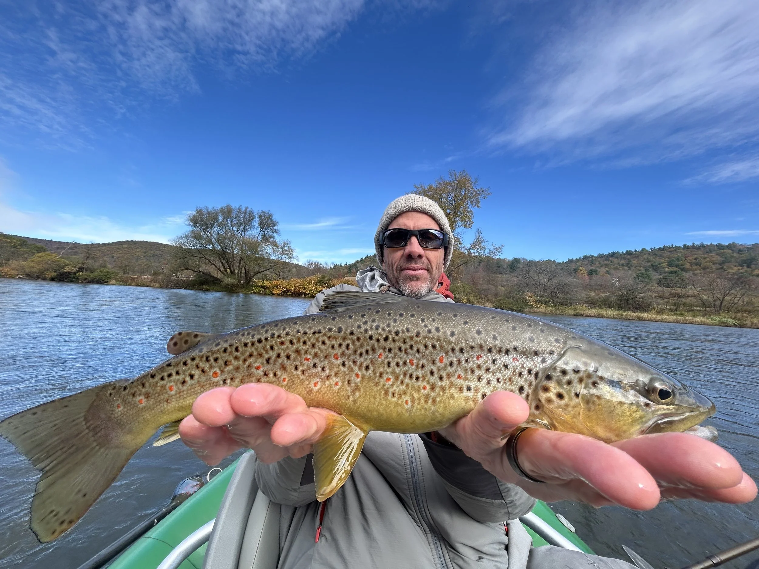 A man wearing sunglasses, a gray beanie, and a gray jacket holding a large brown trout in a boat on a lake. The background shows trees, hills, and a partly cloudy sky.