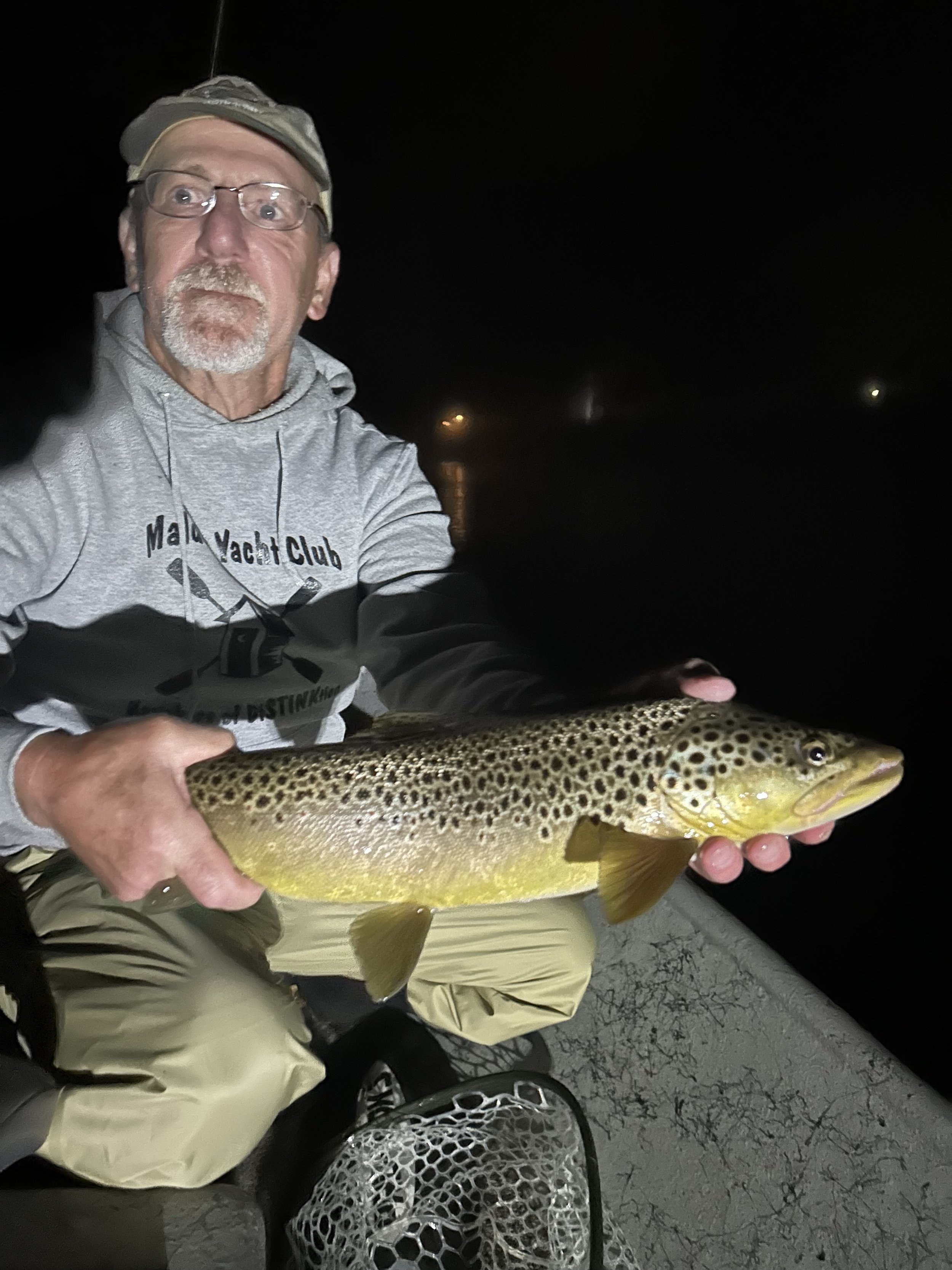 Man holding a large brown trout at night on a boat.