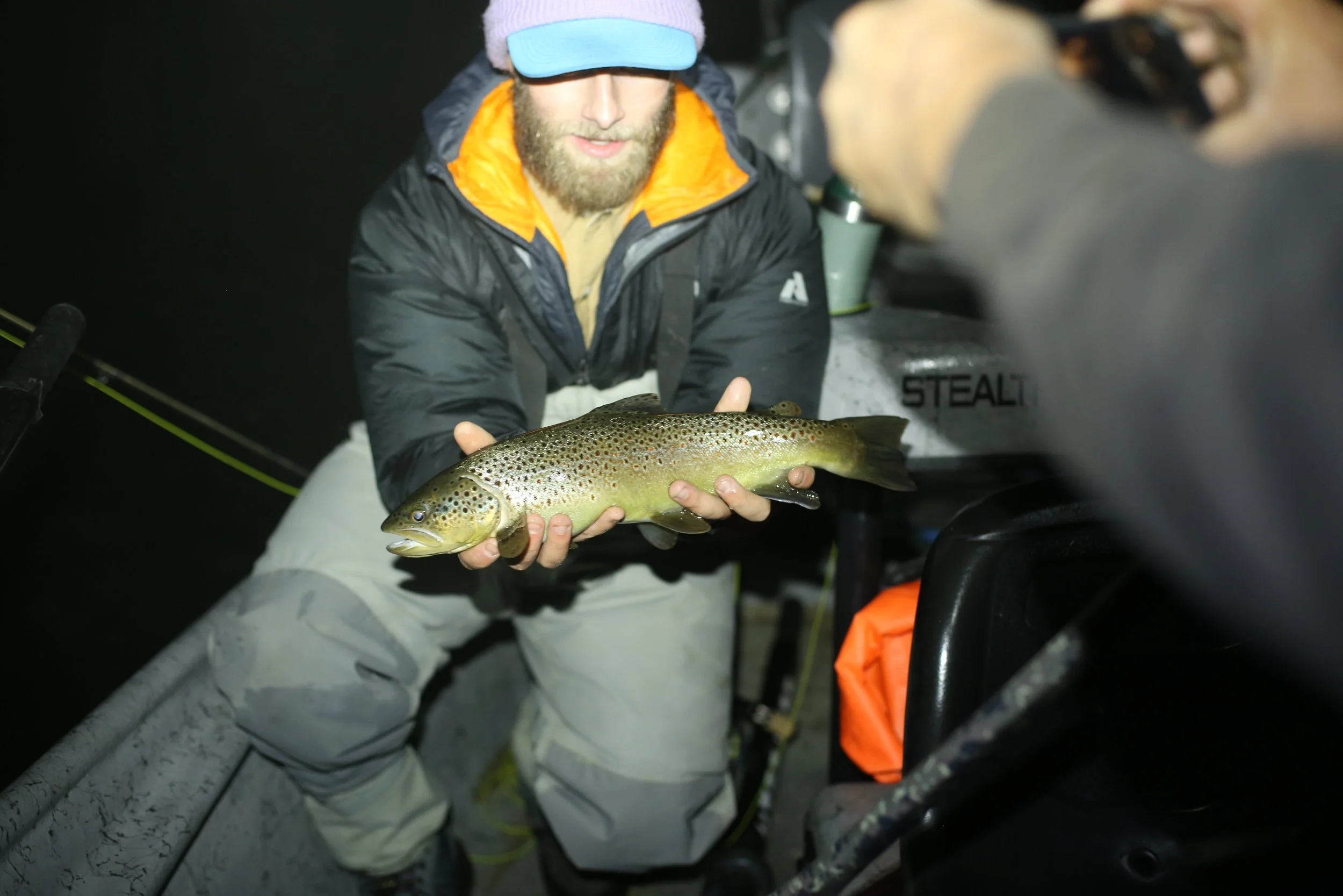 Man holding a large brown trout fish on a boat at night, wearing a pink beanie, sunglasses, and outdoor clothing.
