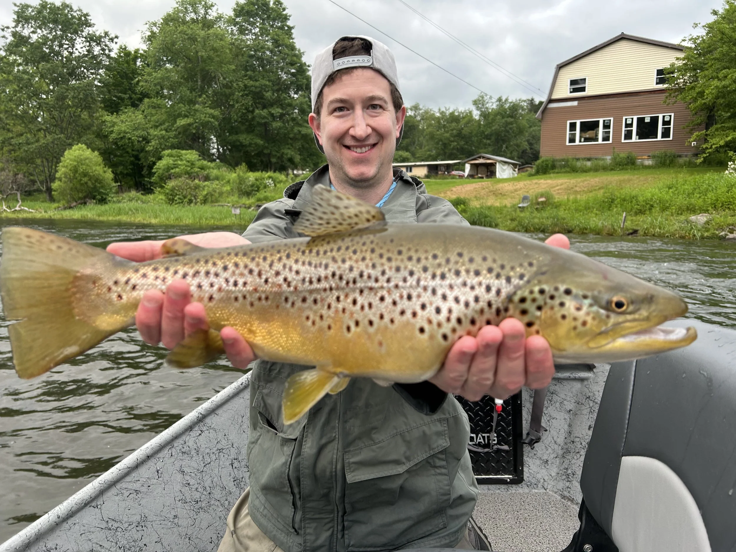 A smiling man in a gray jacket and baseball cap holding a large brown trout fish on a boat in a river with green trees and houses in the background.