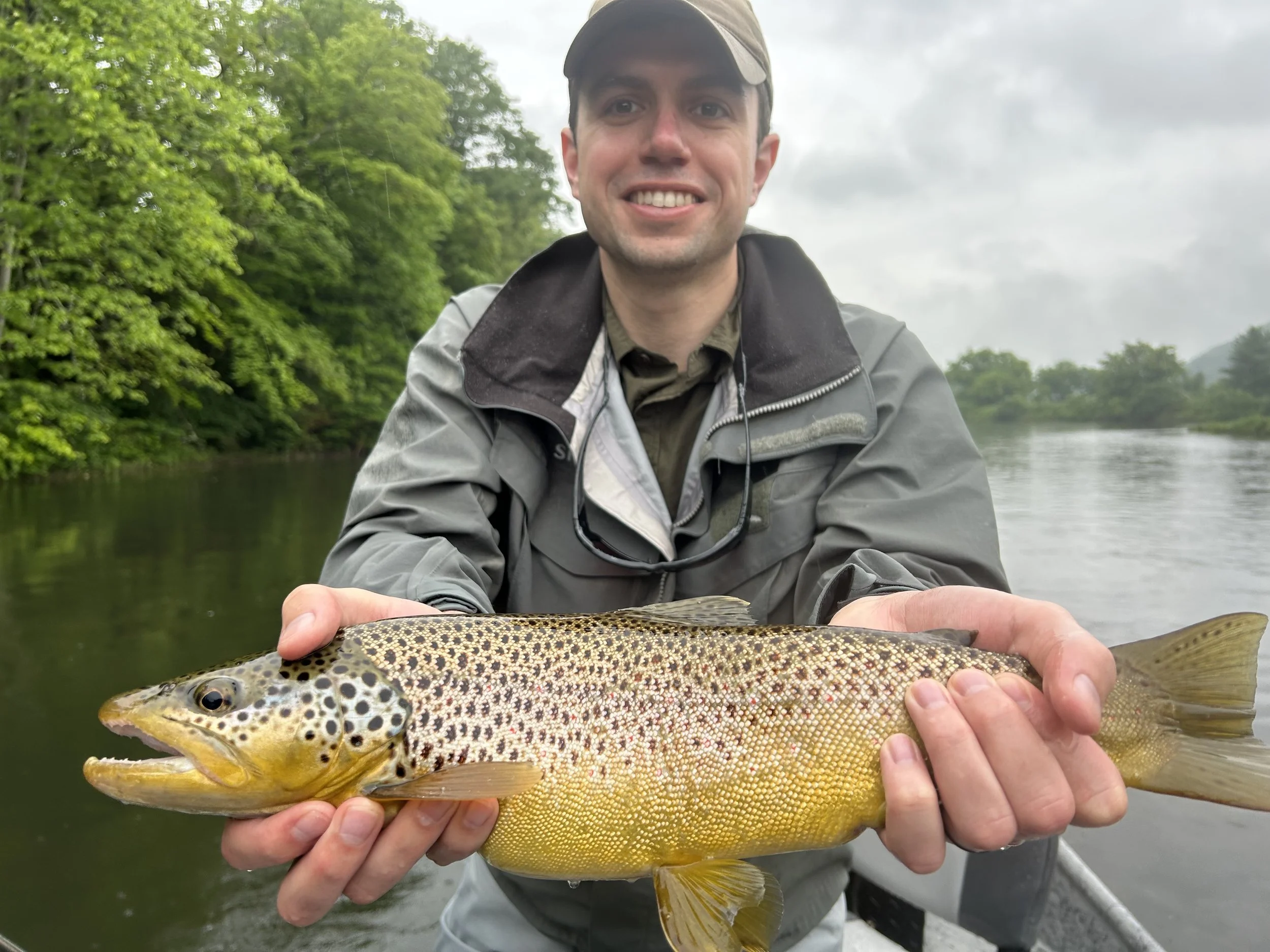 Man in outdoor gear smiling while holding a large rainbow trout fish on a river.