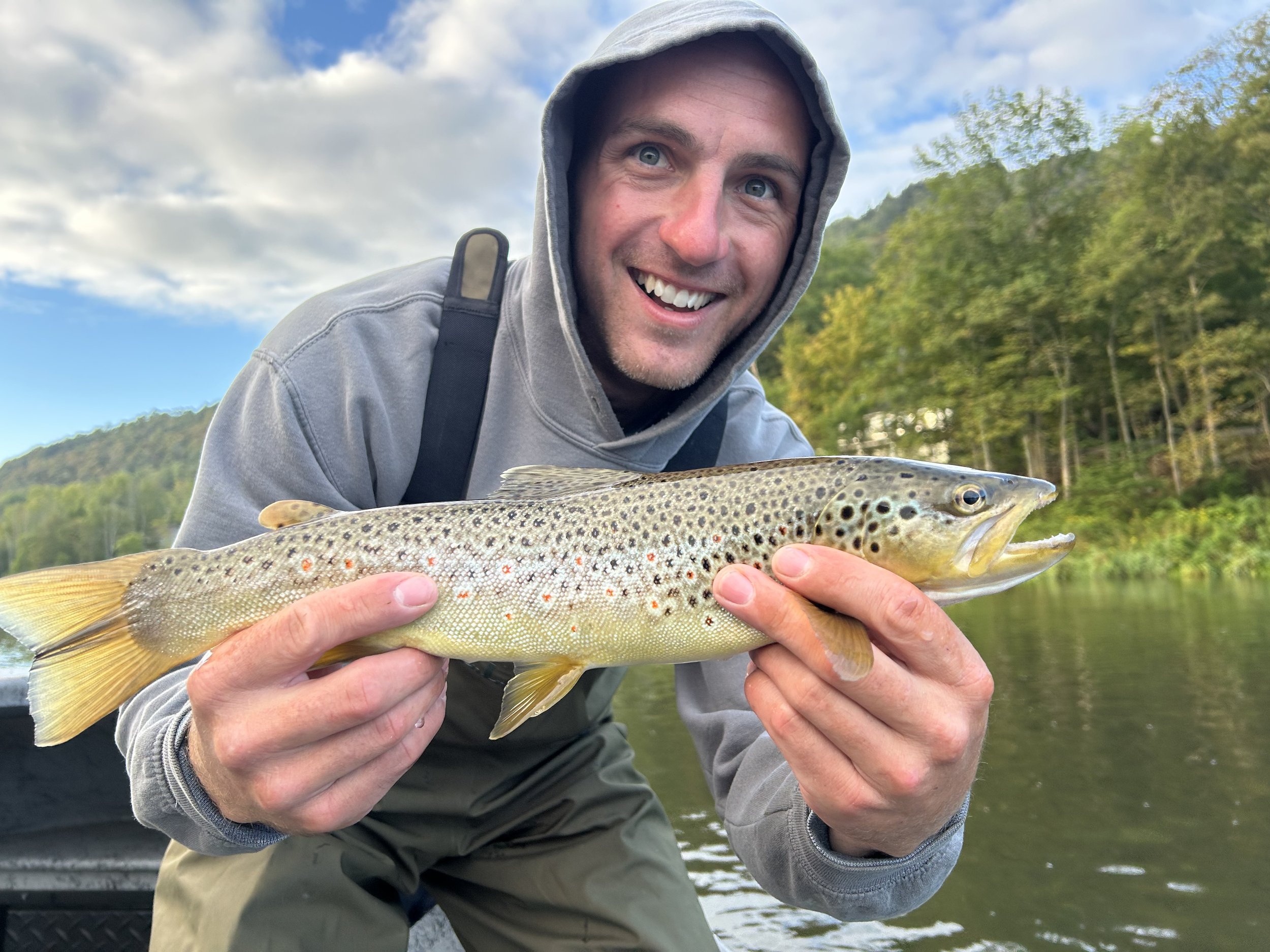 A smiling man wearing a gray hoodie and a backpack holding a large trout fish outdoors near a river, with trees and a cloudy sky in the background.