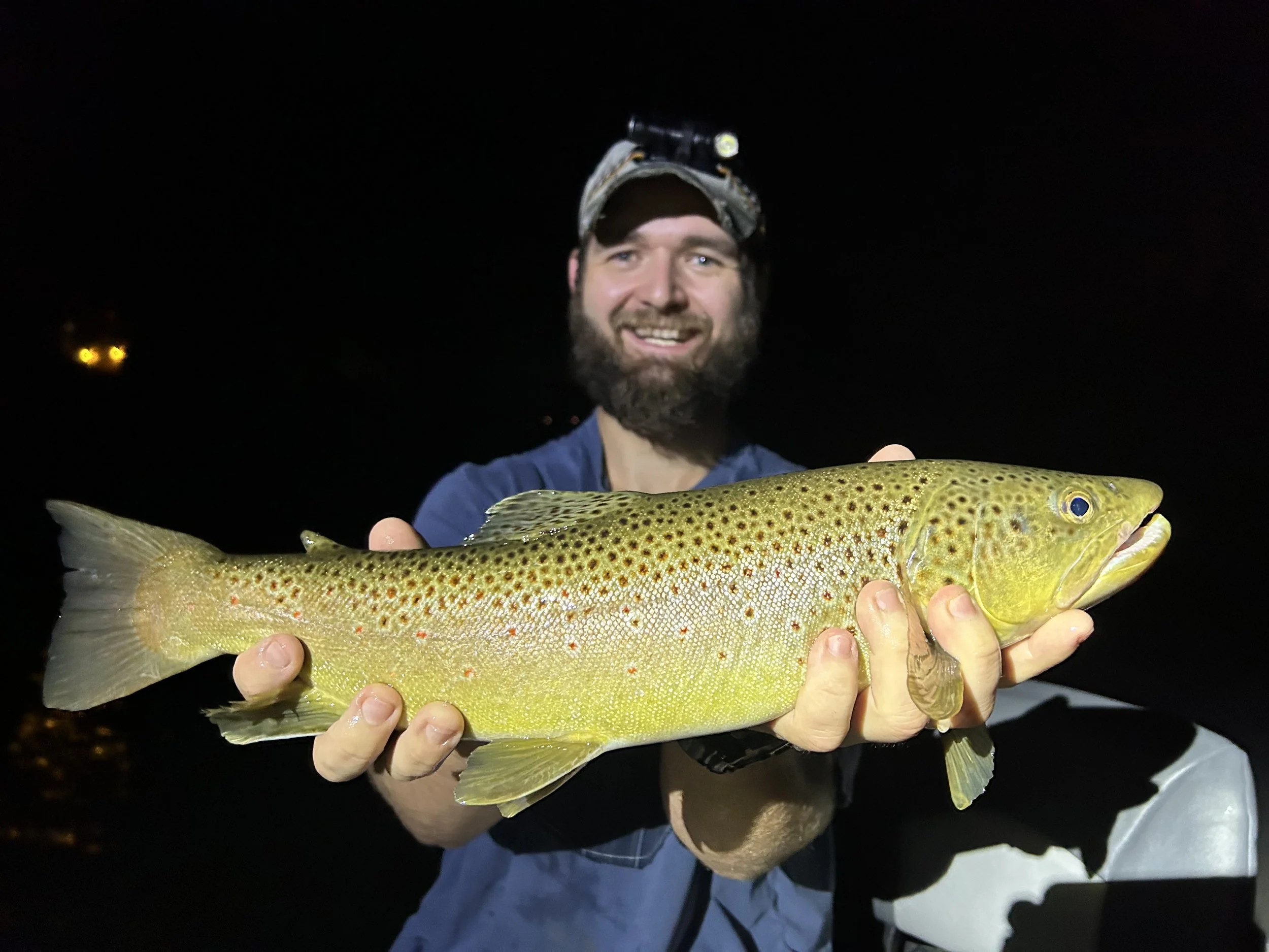 Man holding a large trout fish at night, wearing a headlamp.