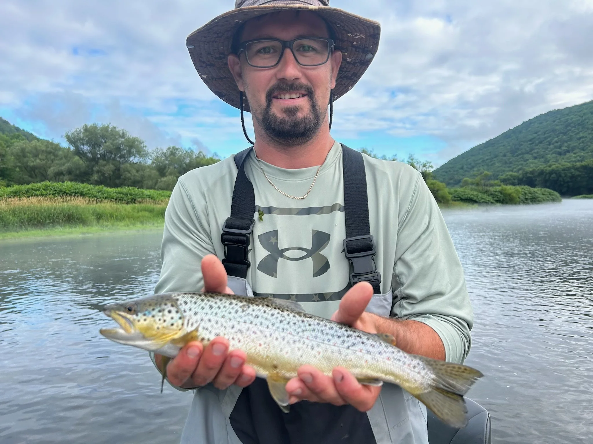 A man wearing a wide-brimmed hat, glasses, and a gray shirt, holding a fish with both hands, standing in front of a river with lush green trees and hills in the background.