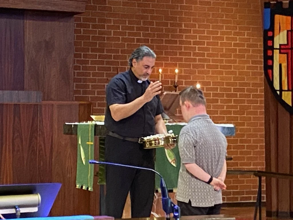 Rev. Nader Hanna baptizing a person standing in front of a church pulpit. Rev. Nader holds a chalice and raises a hand, with a brick wall and lit candles in the background.