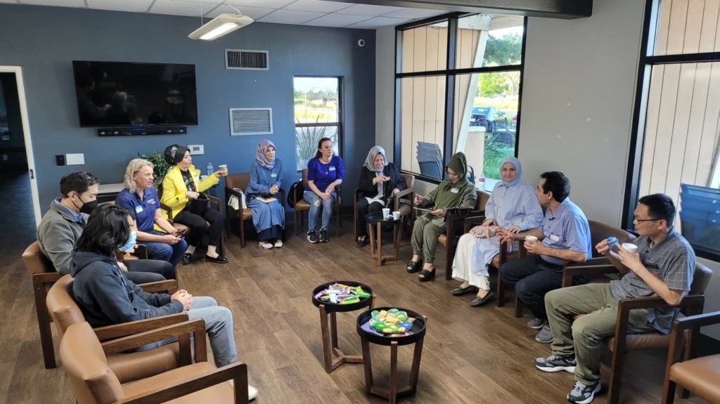 A diverse group of people sitting in a circle in an indoor room, engaging in a discussion or meeting. The room has large windows and a TV on the wall, with snacks on a small central table.